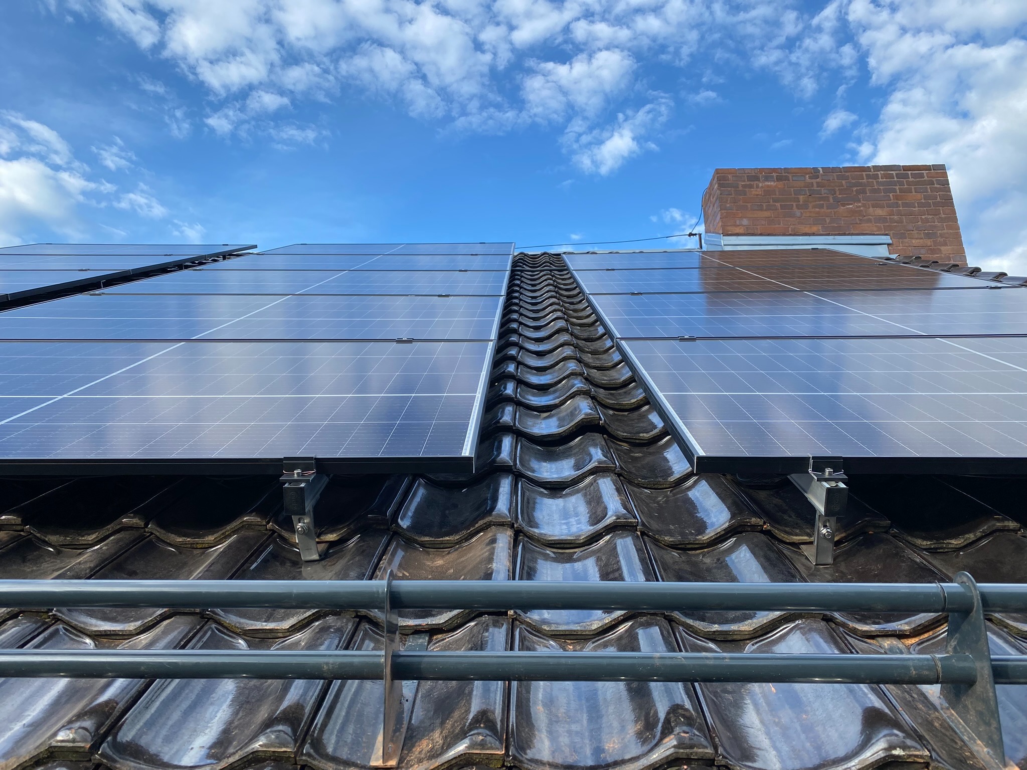 Panele słoneczne zamontowane na błyszczącym, ciemnym dachu z dachówki ceramicznej, widok z dołu na tle błękitnego nieba z białymi chmurami, widoczna drabina zabezpieczająca.
