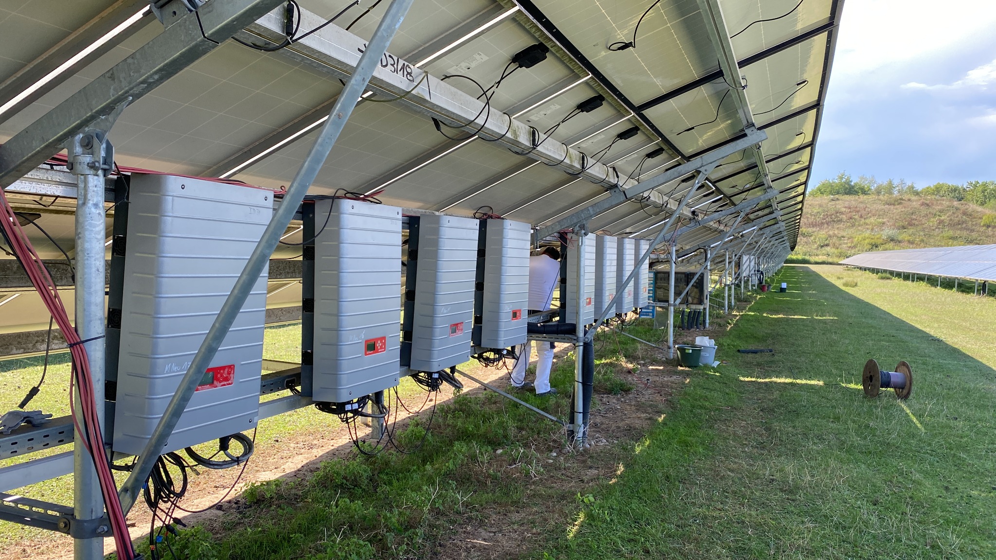 Rząd szarych falowników zamontowanych pod panelami słonecznymi na metalowej konstrukcji, widoczny pracownik w białym kombinezonie podczas instalacji, w tle trawa i kolejne rzędy paneli.