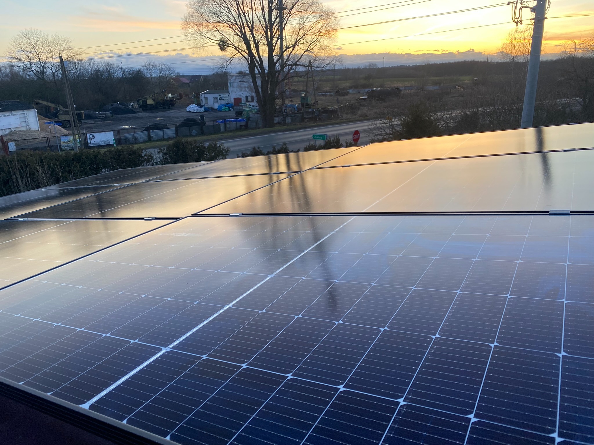 Instalacja fotowoltaiczna na dachu budynku, odbijająca zachodzące słońce i okoliczny krajobraz z drzewami i zabudowaniami.