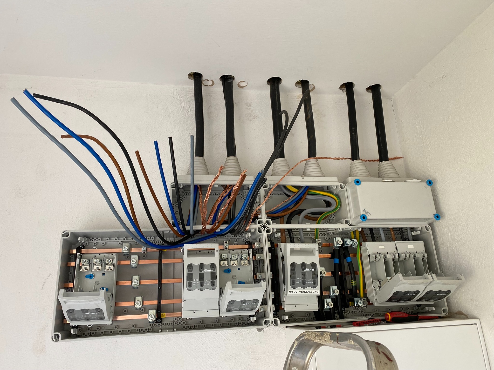 Otwarta skrzynka rozdzielcza z widocznymi bezpiecznikami i przewodami elektrycznymi w różnych kolorach, zamontowana na białej ścianie. Widoczne narzędzia, drabina i rury w suficie.