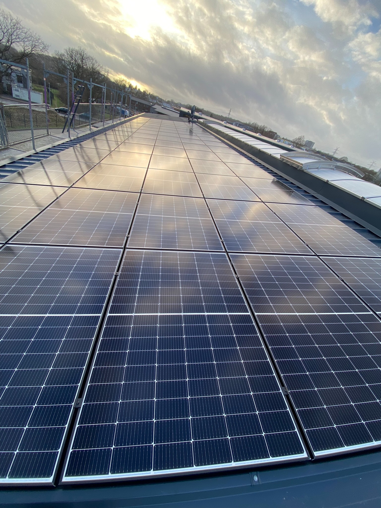 Rząd paneli słonecznych na dachu budynku odbijający zachmurzone niebo, z widocznym fragmentem otoczenia i sylwetką osoby w oddali.