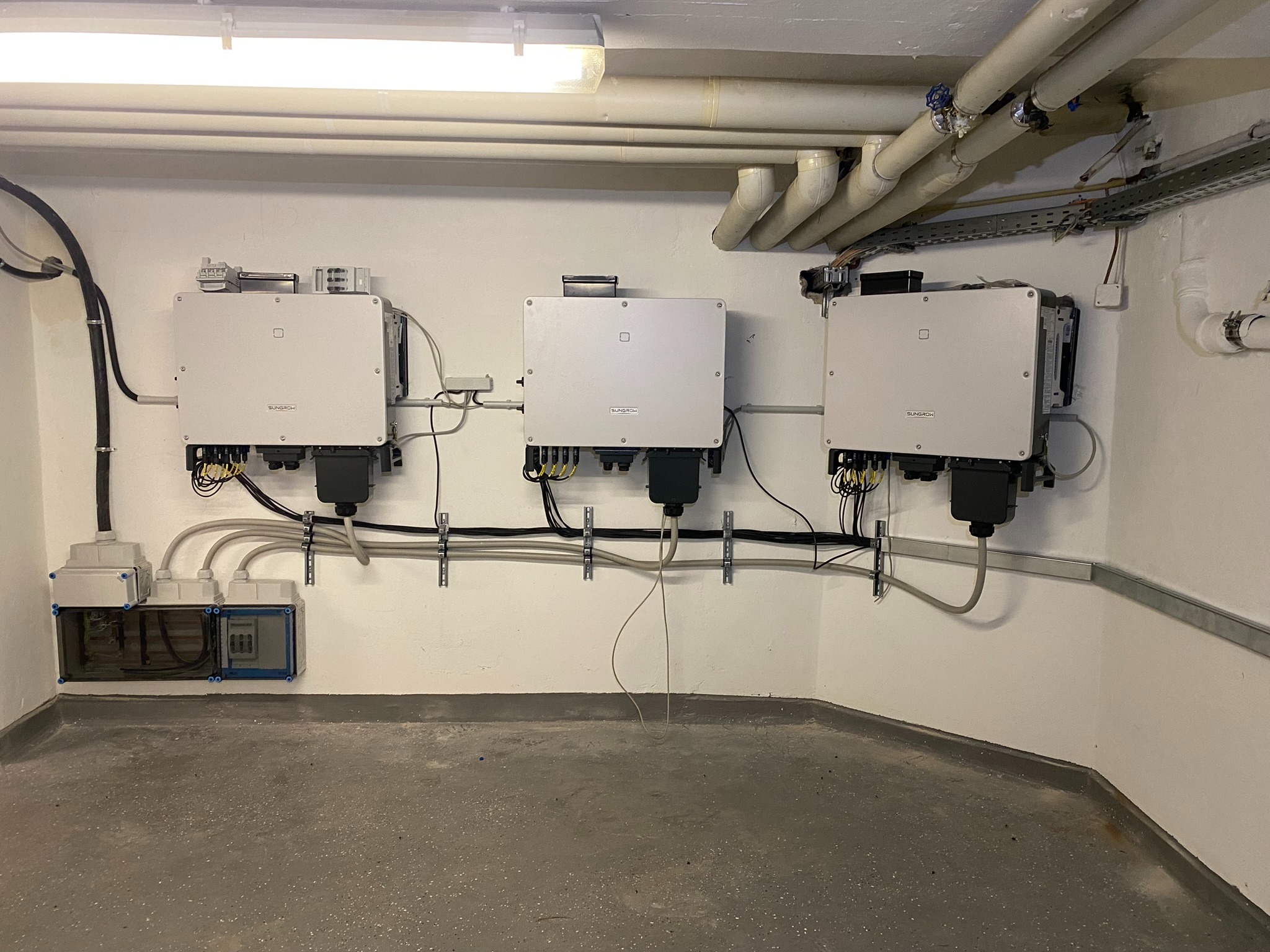 Trzy białe falowniki marki Sungrow zamontowane na ścianie w pomieszczeniu technicznym, widoczne kable i rury instalacyjne pod sufitem.