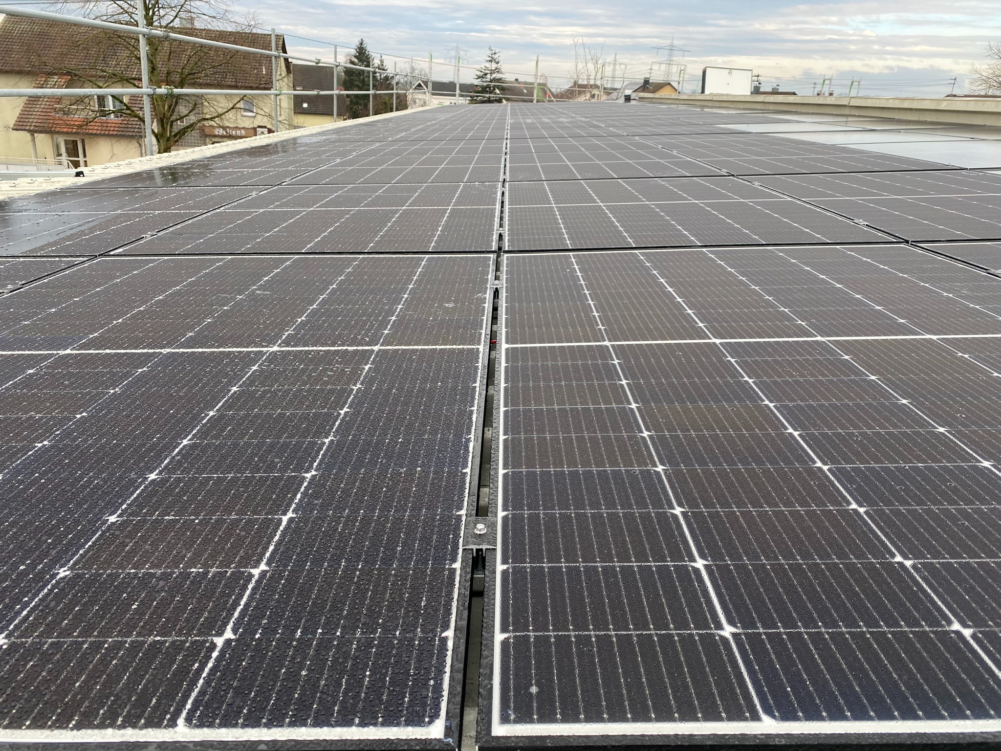 Widok z bliska na mokre panele słoneczne na dachu budynku w pochmurny dzień, widoczne elementy montażowe i otoczenie miejskie.