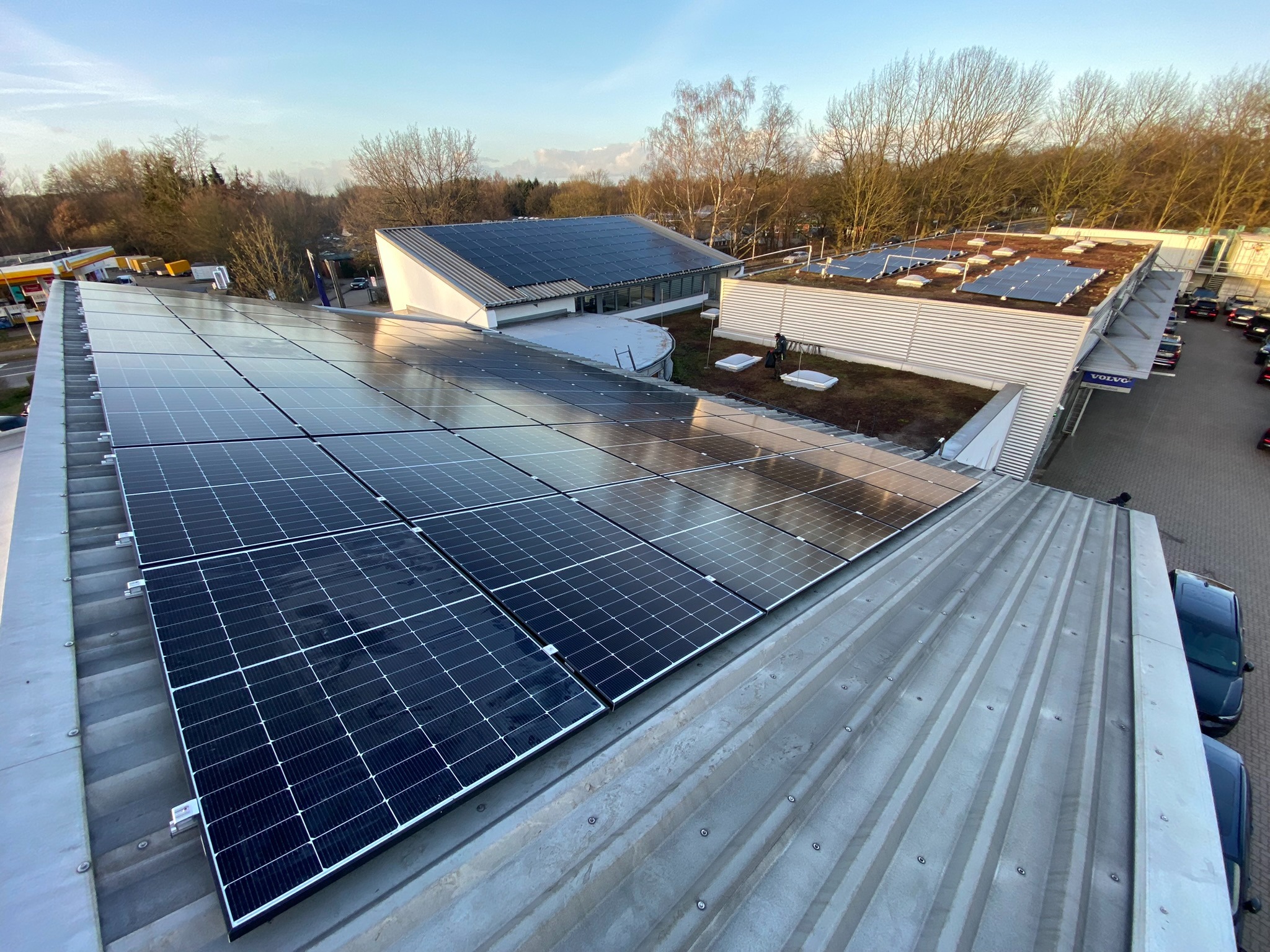 Instalacja paneli słonecznych na dachach budynków o różnej architekturze, w tle drzewa i błękitne niebo.