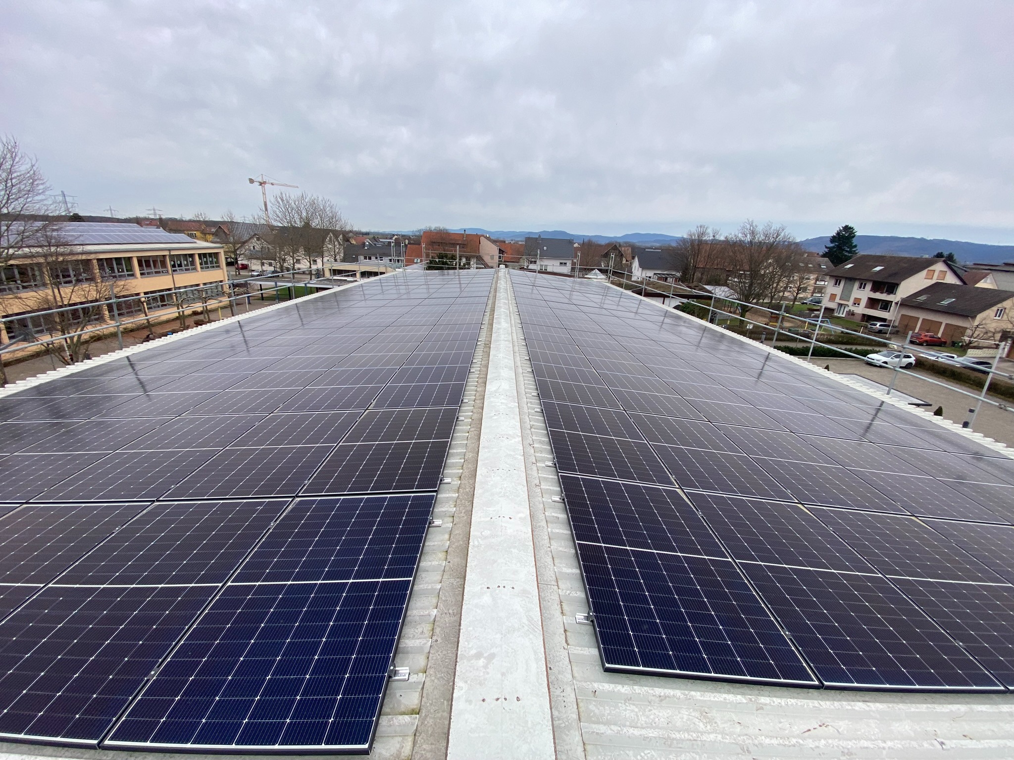 Rozległa instalacja paneli słonecznych na dachu budynku, widok z góry, w tle zabudowa miejska i pochmurne niebo.