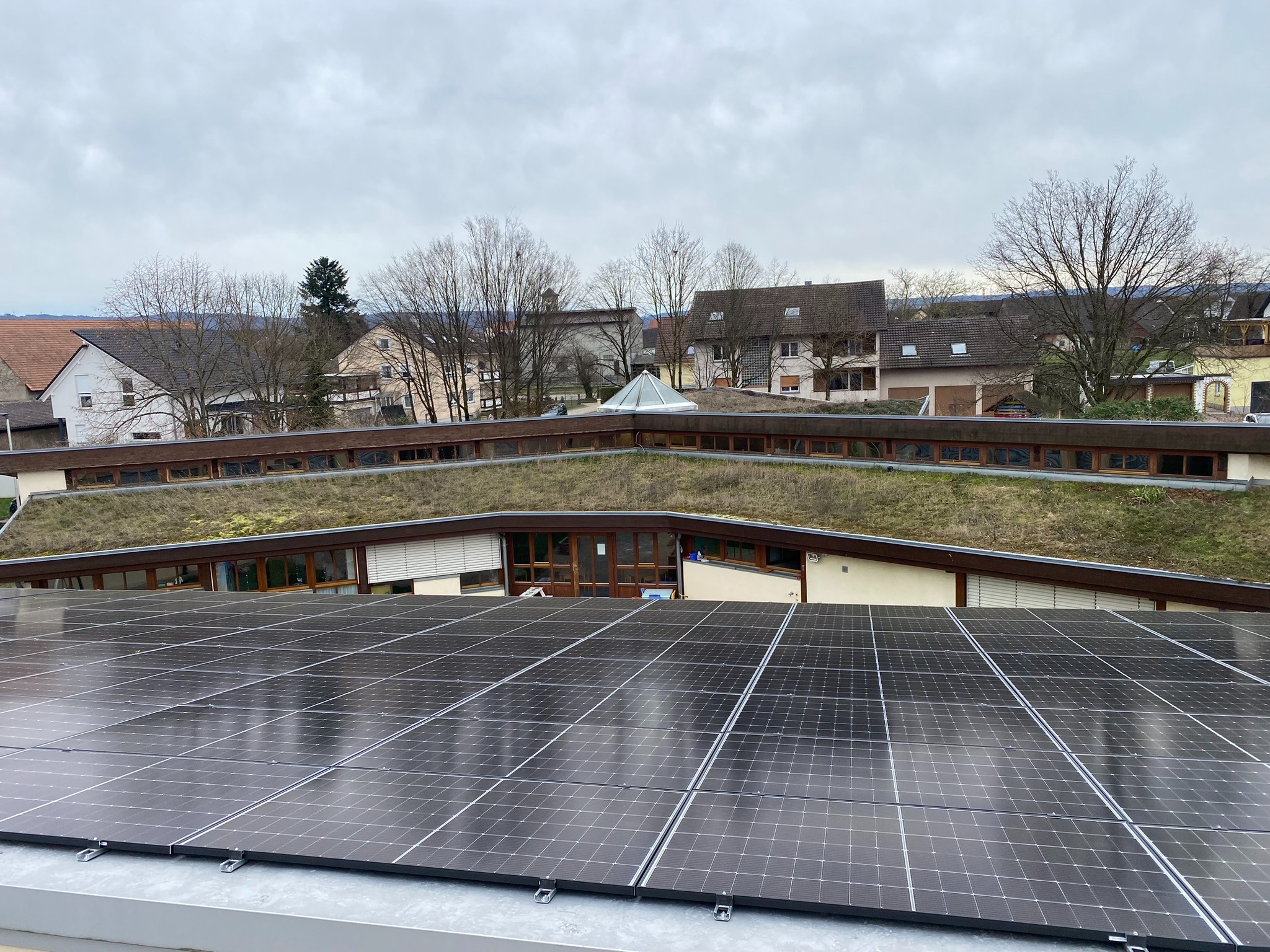 Instalacja fotowoltaiczna na płaskim dachu budynku z zielonym dachem w tle zabudowy jednorodzinnej pod pochmurnym niebem.