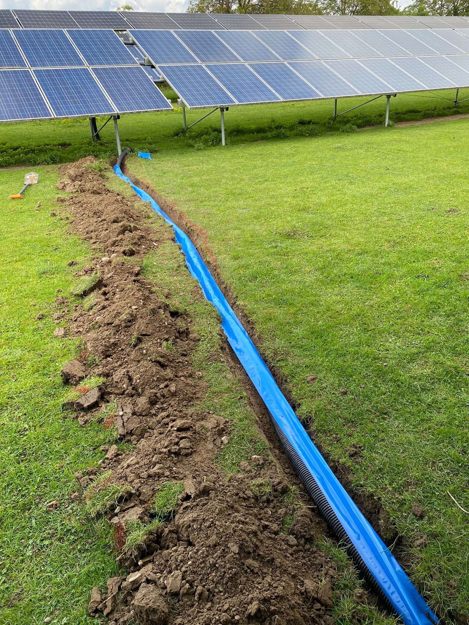 Wykop z niebieską folią ochronną i czarnym karbowanym przewodem, prawdopodobnie do instalacji elektrycznej, na tle paneli słonecznych na trawiastym terenie.