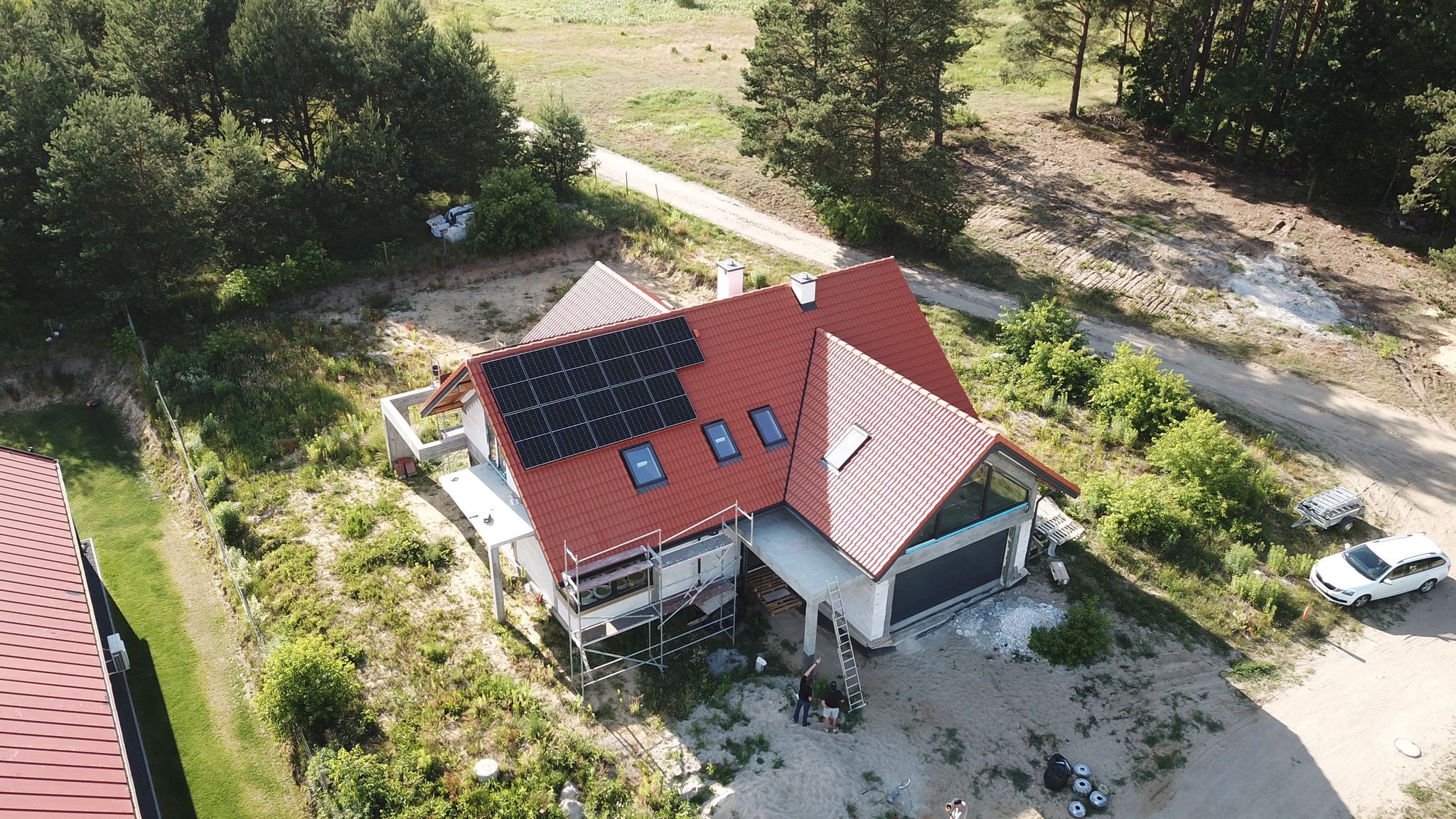 Widok z drona na dom jednorodzinny z czerwoną dachówką i panelami słonecznymi na dachu, częściowo otoczony rusztowaniami, z widocznymi pracownikami na podwórku przed garażem i samochodem osobowym...