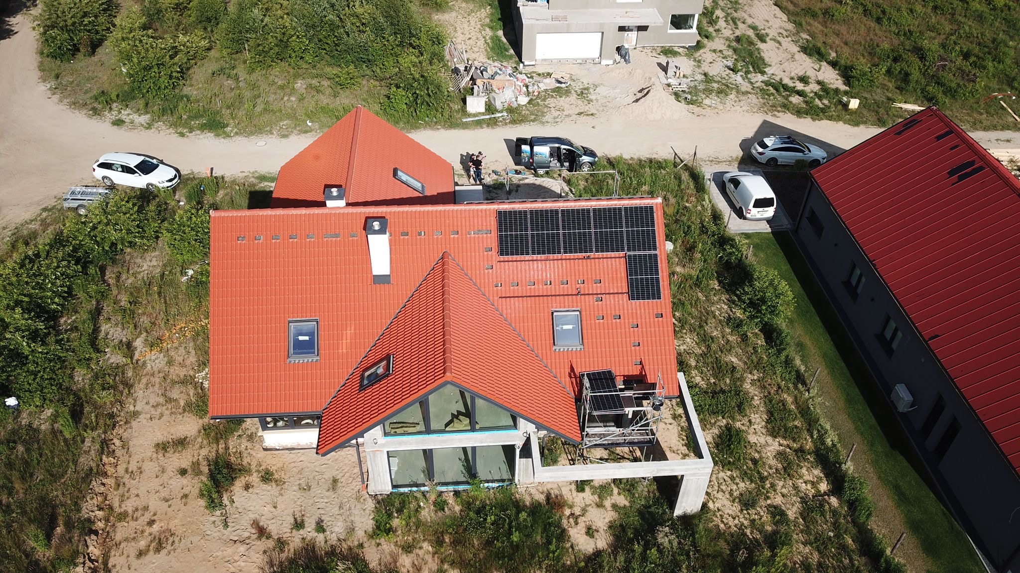 Widok z lotu ptaka na dom z czerwoną dachówką i panelami słonecznymi w trakcie montażu, widoczne rusztowanie i ekipa montażowa przy samochodzie dostawczym.