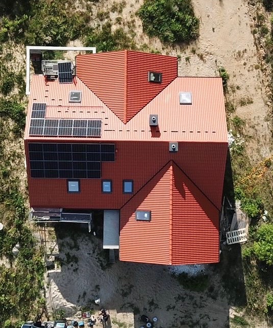 Widok z góry na dom z czerwoną dachówką i panelami słonecznymi, kilka okien dachowych, otoczony zielenią i piaskiem.