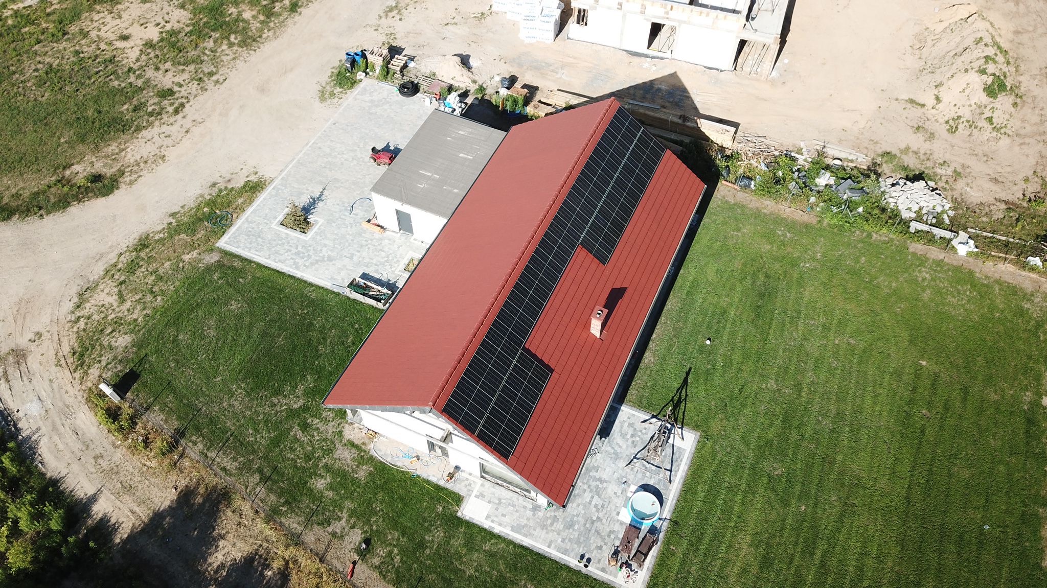 Widok z drona na dom z czerwoną dachówką i zamontowanymi panelami słonecznymi. Na trawniku obok domu stoi huśtawka i jacuzzi. W tle widać budowę innego domu.