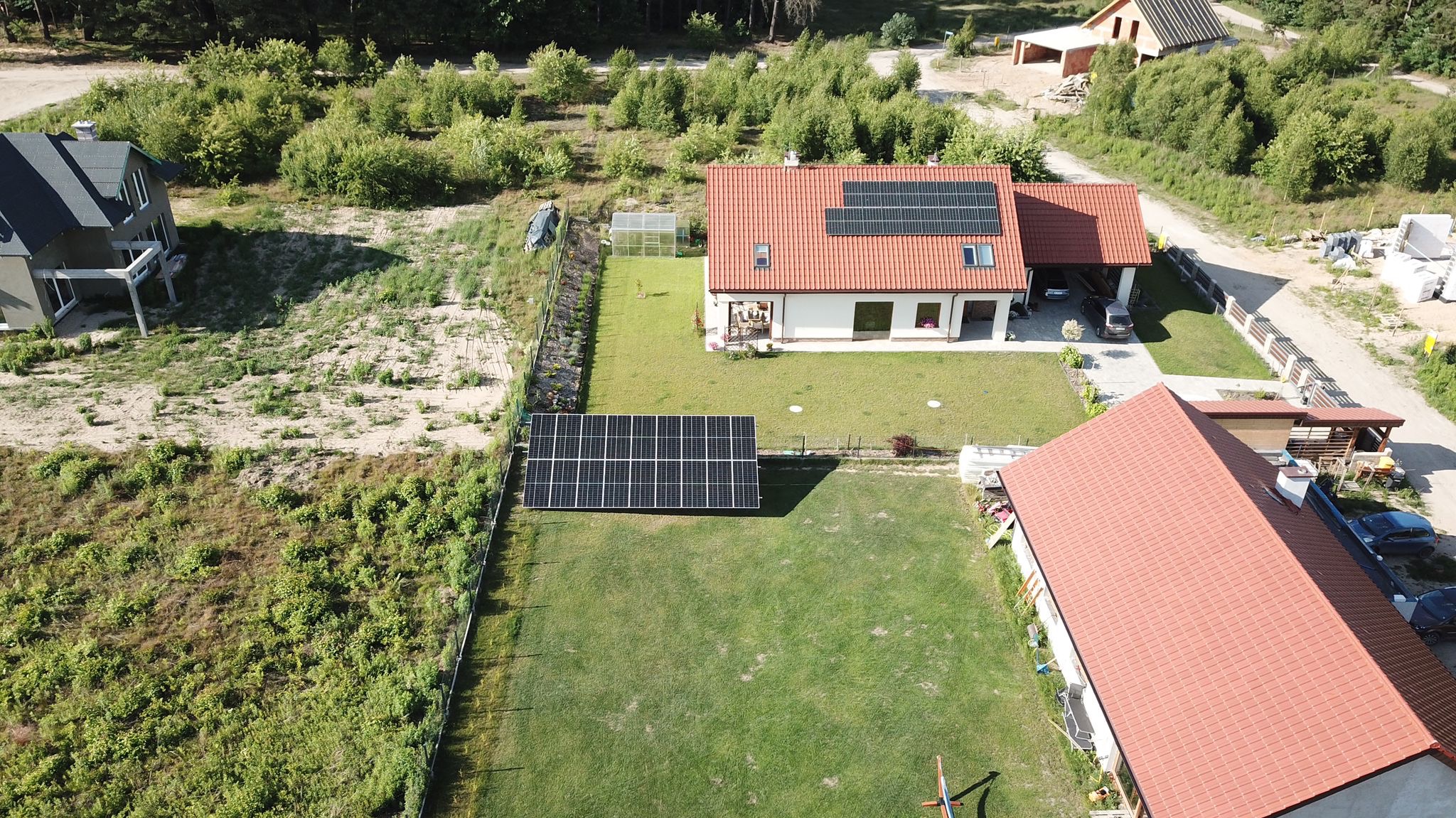 Widok z drona na dom jednorodzinny z panelami słonecznymi na dachu i wolnostojącą instalacją paneli na trawniku, otoczony zielenią i innymi budynkami.
