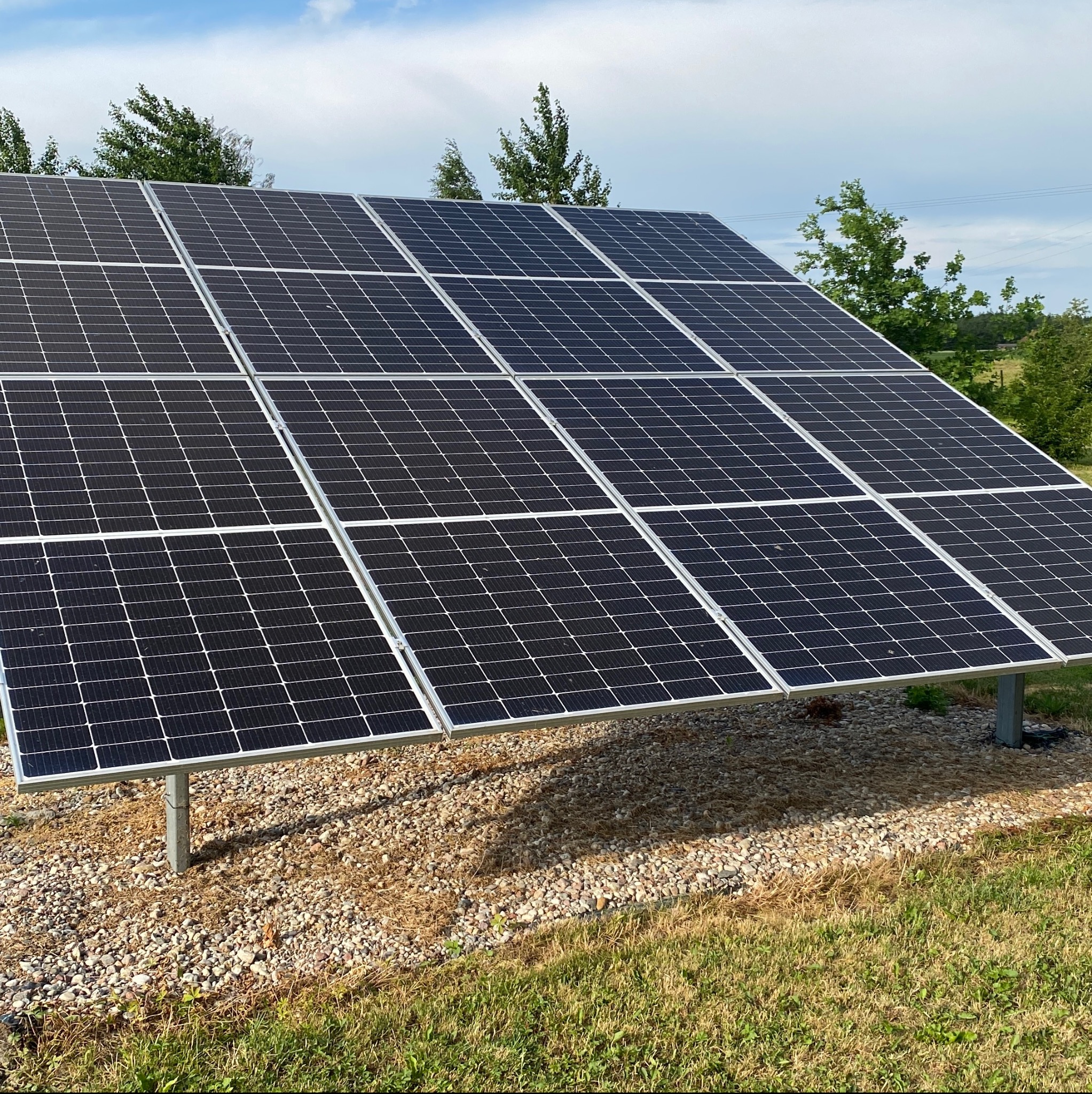 Instalacja fotowoltaiczna składająca się z kilkunastu paneli na metalowych podporach, otoczona żwirem i trawą, częściowo zacieniona.