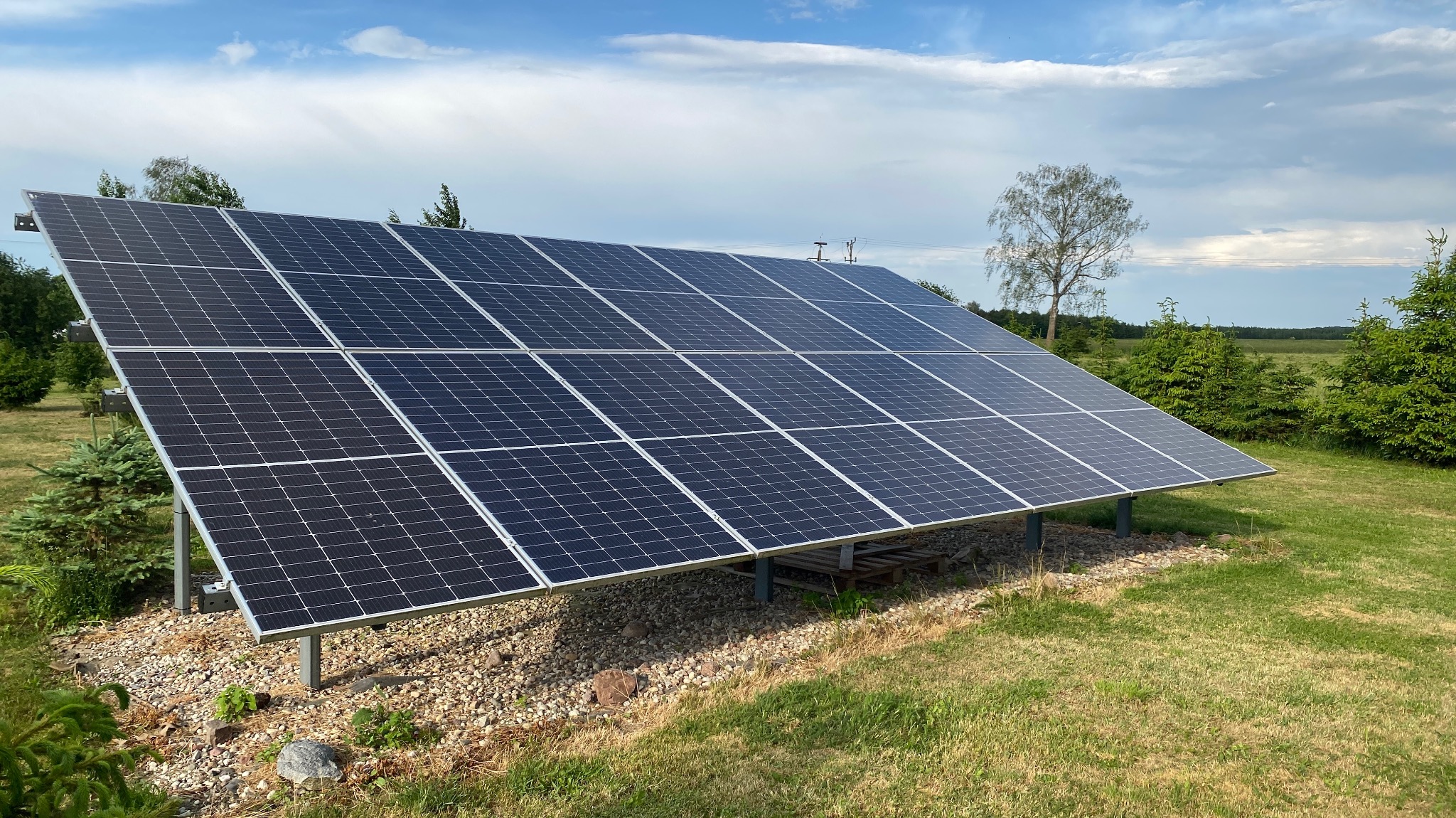 Instalacja paneli słonecznych na metalowej konstrukcji, widoczna na tle zielonego trawnika i drzew, z niebem w tle.