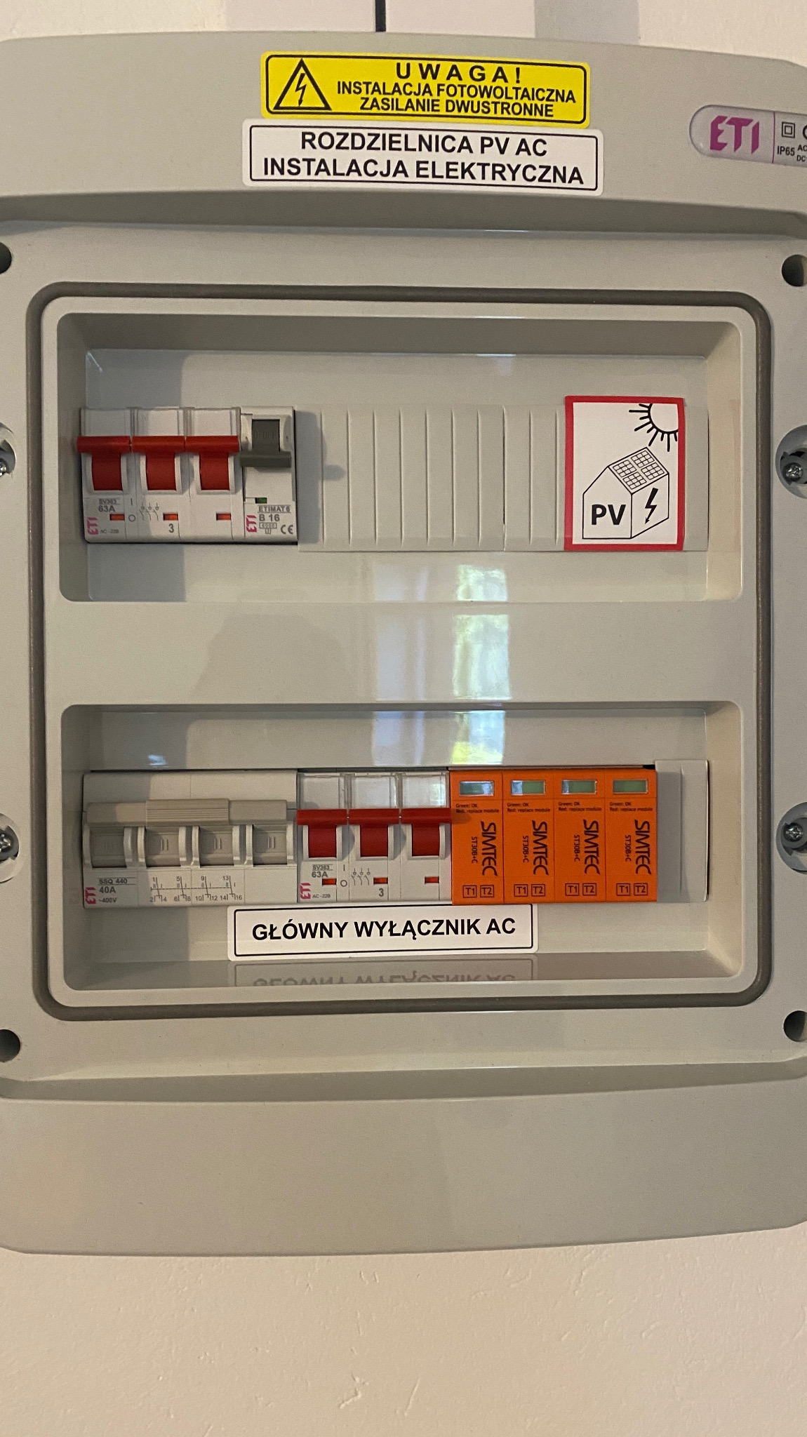Rozdzielnica PV AC z wyłącznikami ETI i SIMTEC, naklejka ostrzegawcza o instalacji fotowoltaicznej i zasilaniu dwustronnym, etykieta 'Główny Wyłącznik AC'.