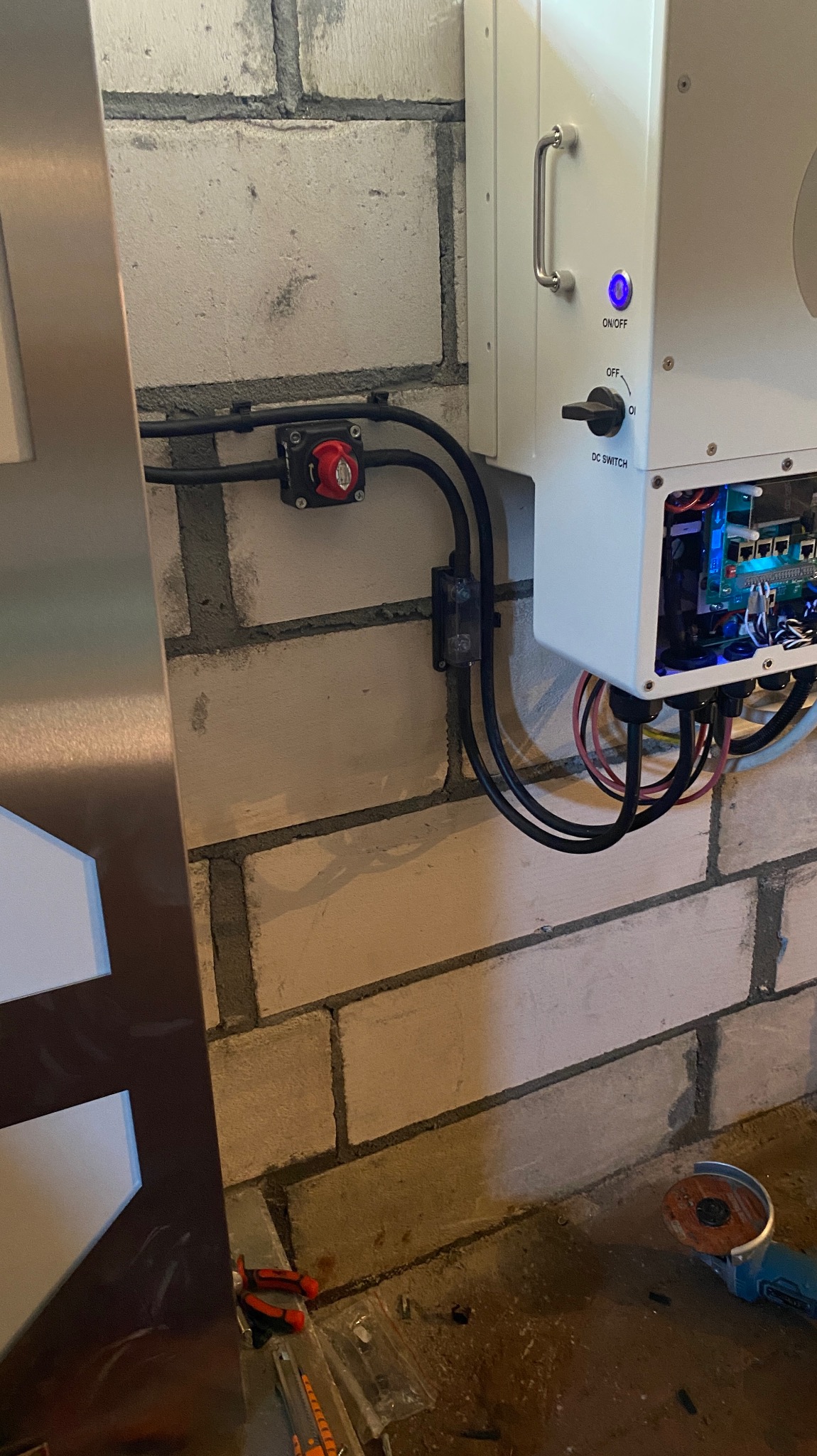Biała obudowa falownika z widocznym wnętrzem i czarnymi kablami przymocowana do ściany z białej cegły, obok wyłącznik bezpieczeństwa. Na podłodze widoczna szlifierka kątowa.
