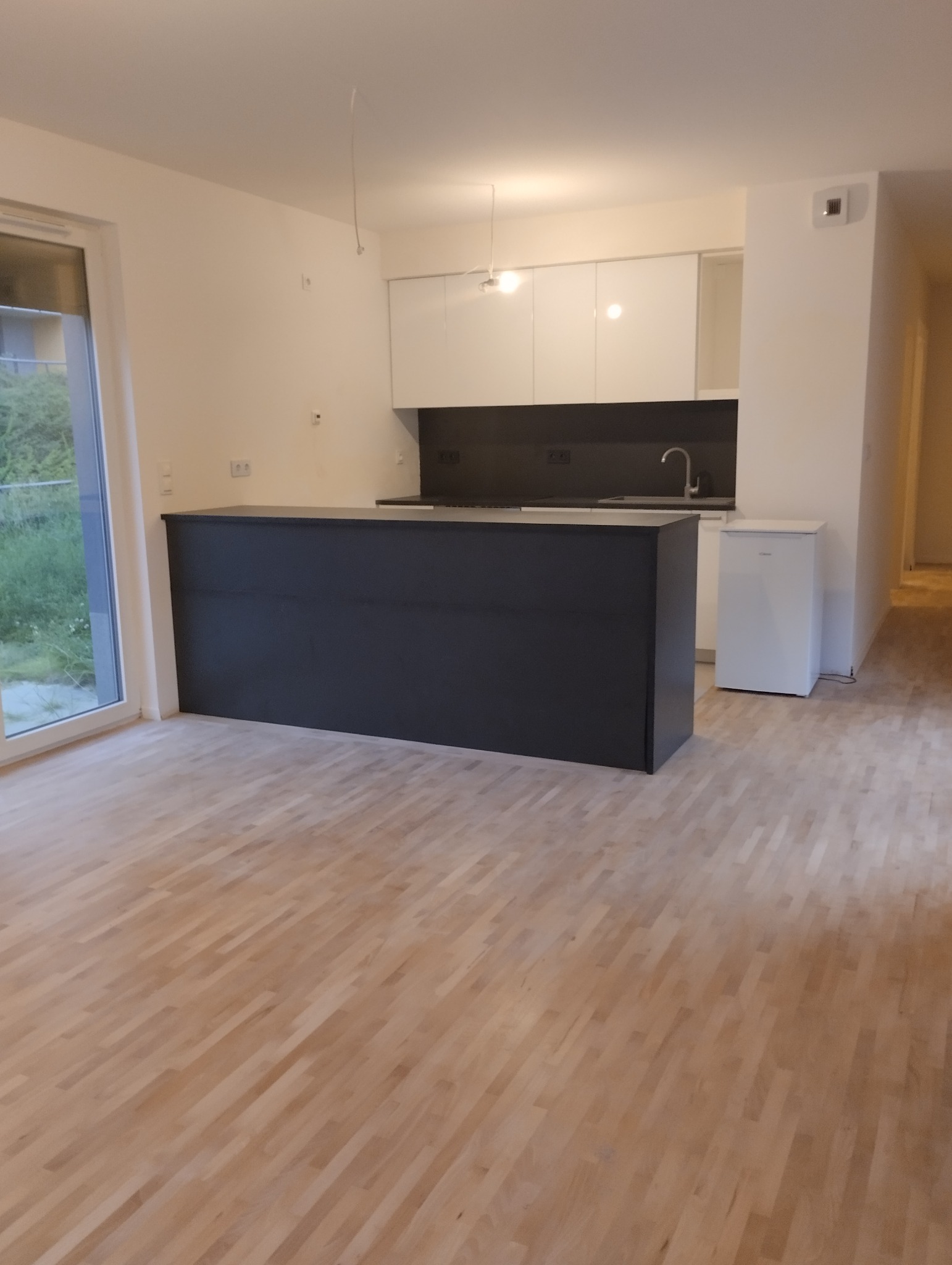 Nowoczesna, jasna kuchnia z ciemnoszarą wyspą kuchenną, białymi szafkami i drewnianą podłogą parkietową ułożoną w jodełkę, widok z perspektywy podłogi.