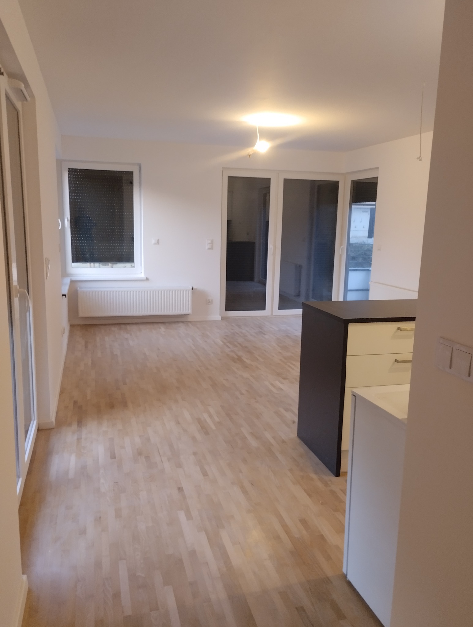 Jasne, puste wnętrze mieszkania z drewnianą podłogą, białymi ścianami, oknem z żaluzjami i zabudową kuchenną z ciemnym blatem.