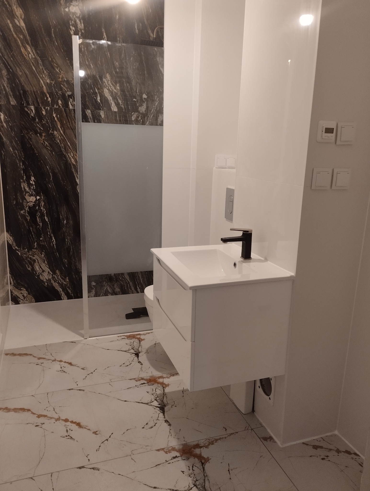 Nowoczesna łazienka z marmurową podłogą i ścianą prysznicową, białą umywalką z czarną baterią oraz minimalistycznymi włącznikami światła.