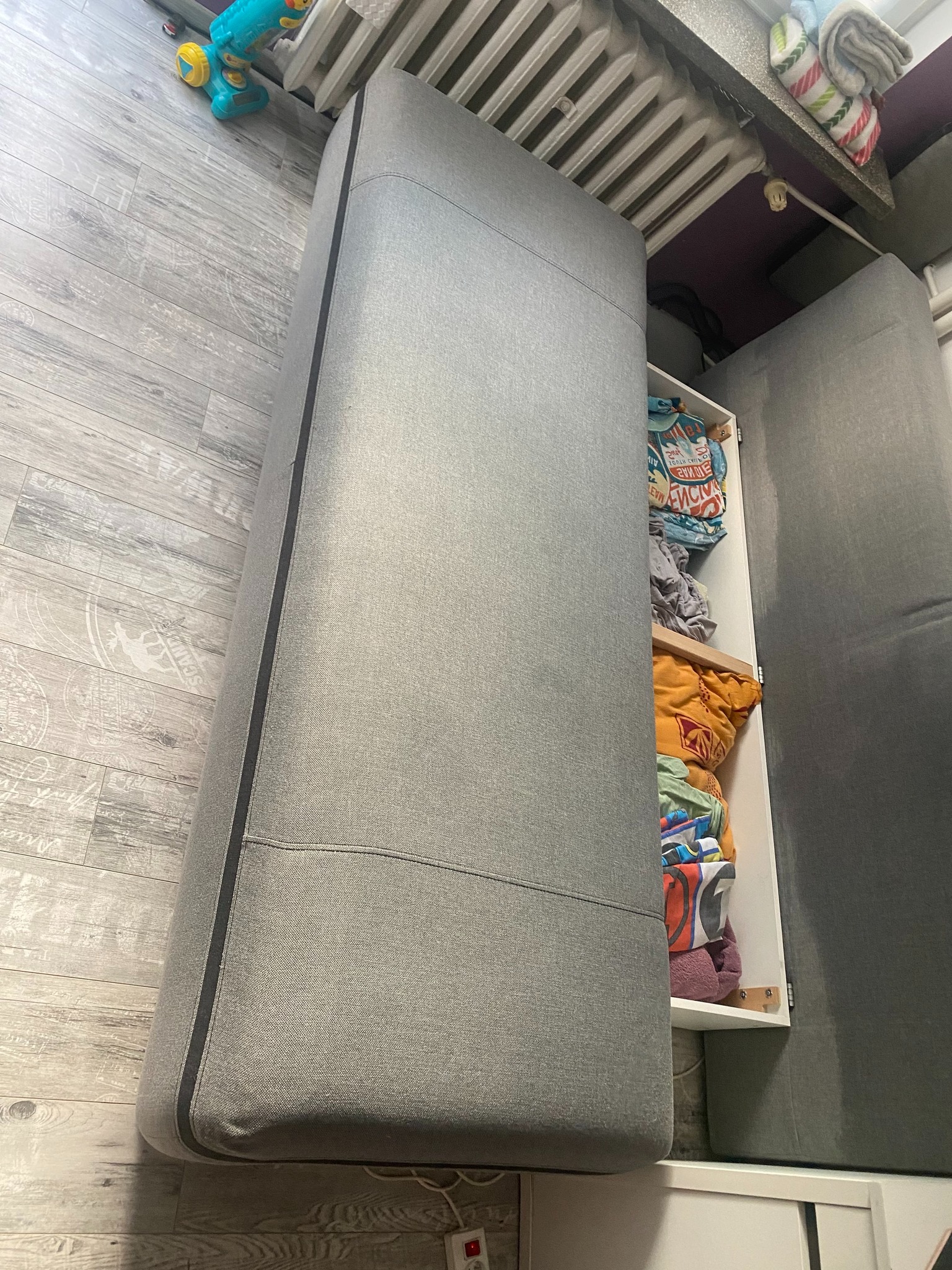 Szara kanapa z wysuwanym pojemnikiem na pościel, wypełnionym ubraniami, na tle szarej podłogi imitującej drewno.