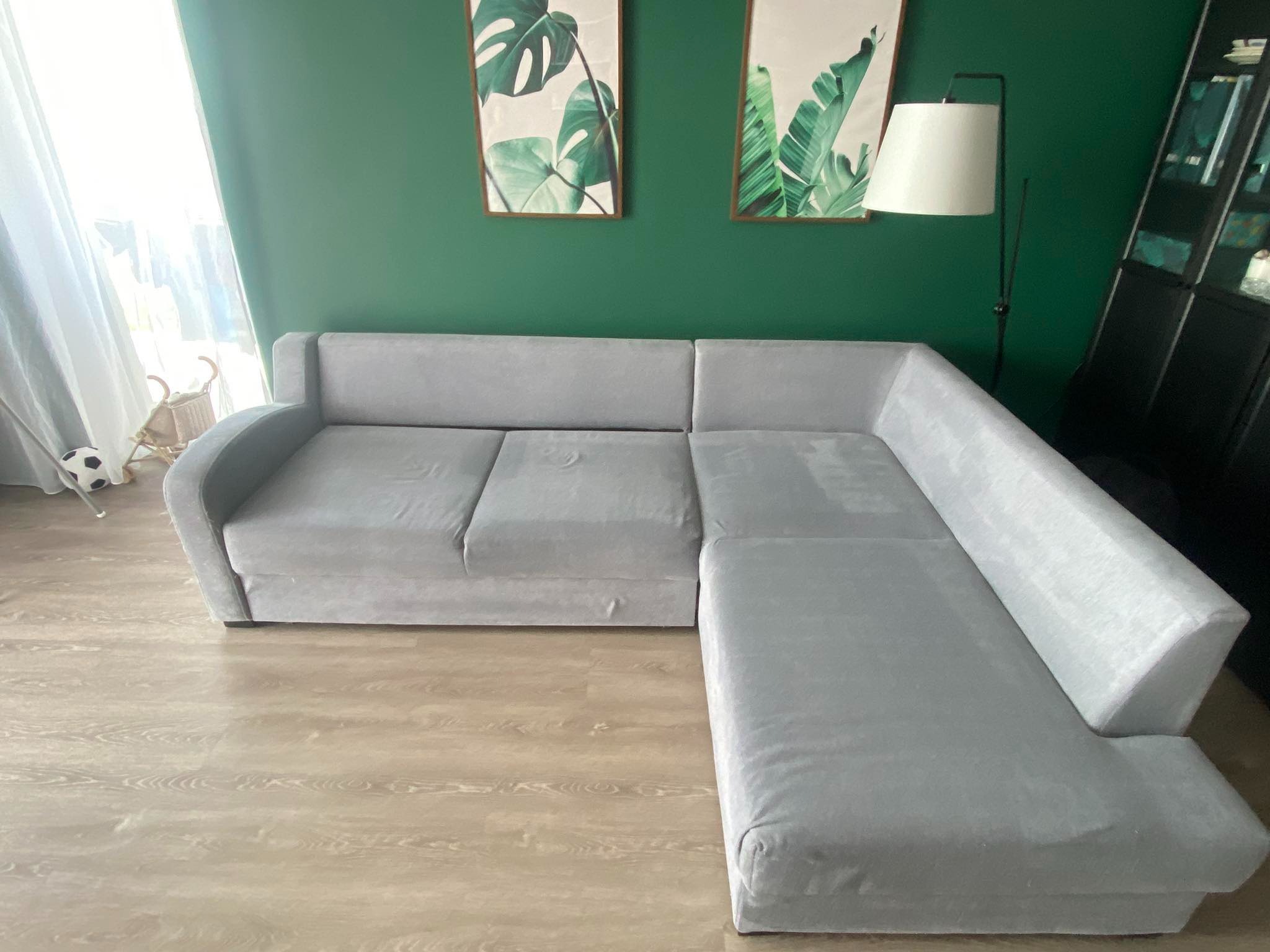 Szara, narożna kanapa w salonie z zieloną ścianą, ozdobiona obrazami z motywami roślinnymi, oświetlona lampą stojącą.