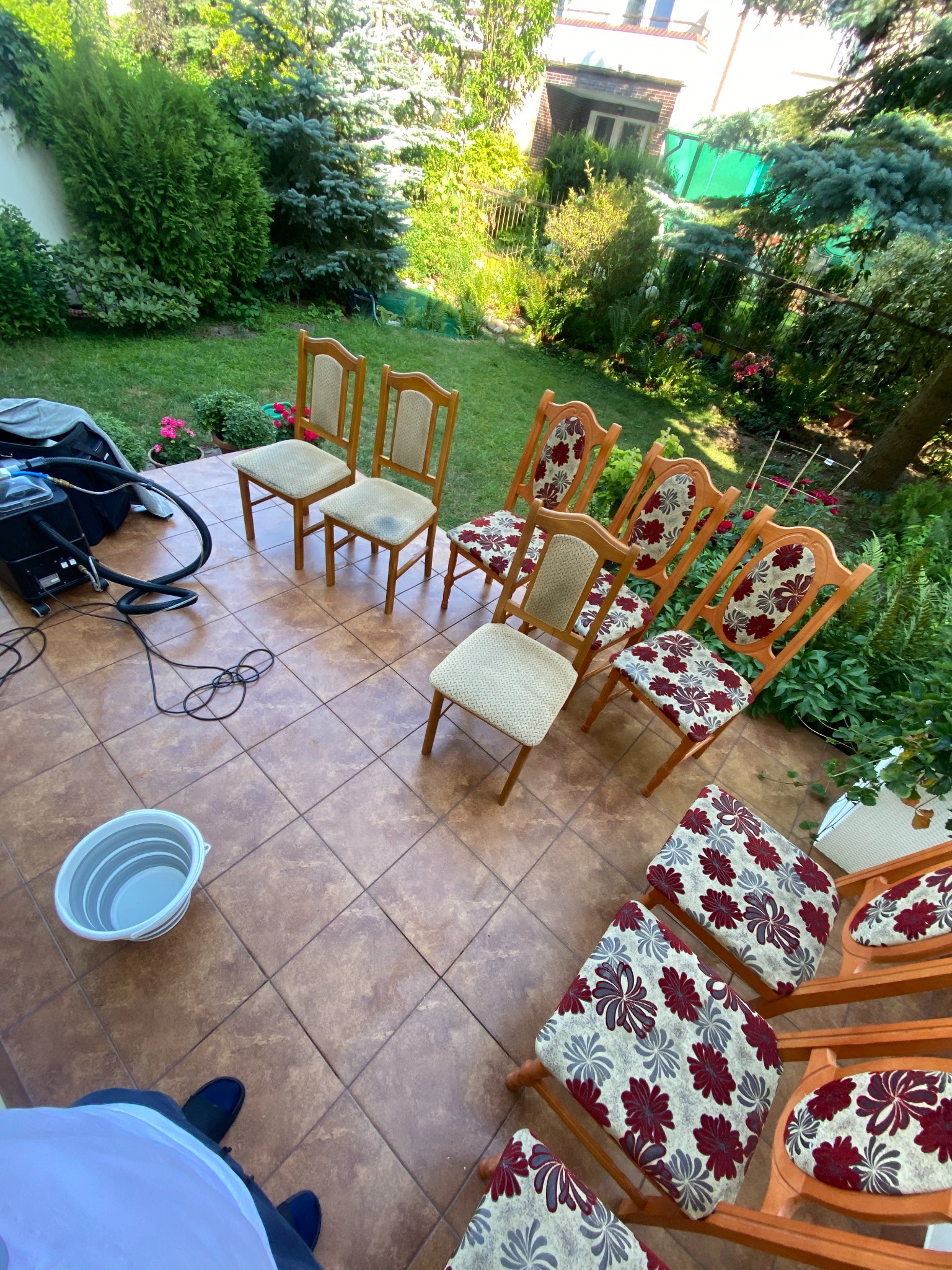 Krzesła ustawione na zewnątrz na kafelkach, obok stoi wiadro i urządzenie do czyszczenia tapicerki, widoczne różne wzory tapicerki krzeseł na tle zieleni ogrodu.