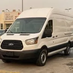 Biały, wysoki furgon Ford Transit zaparkowany na placu, widoczny z boku, z lekko odbijającym się światłem w karoserii. W tle budynek z szyldem w obcym języku i inne pojazdy.