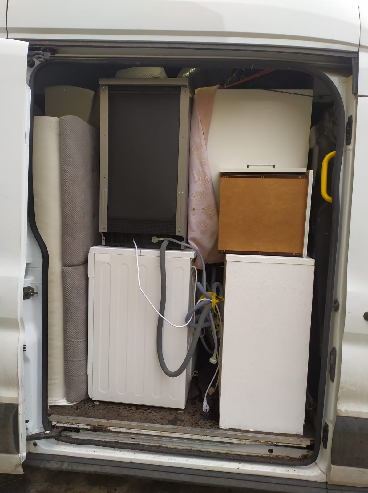 Wnętrze furgonetki załadowane zwiniętymi dywanami, pralką z widocznymi wężami, szafkami oraz innymi przedmiotami podczas transportu.