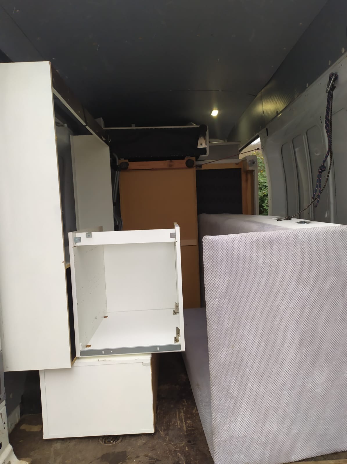 Wnętrze furgonetki załadowane meblami: białe szafki, kartony i materac, zabezpieczone sznurkiem.
