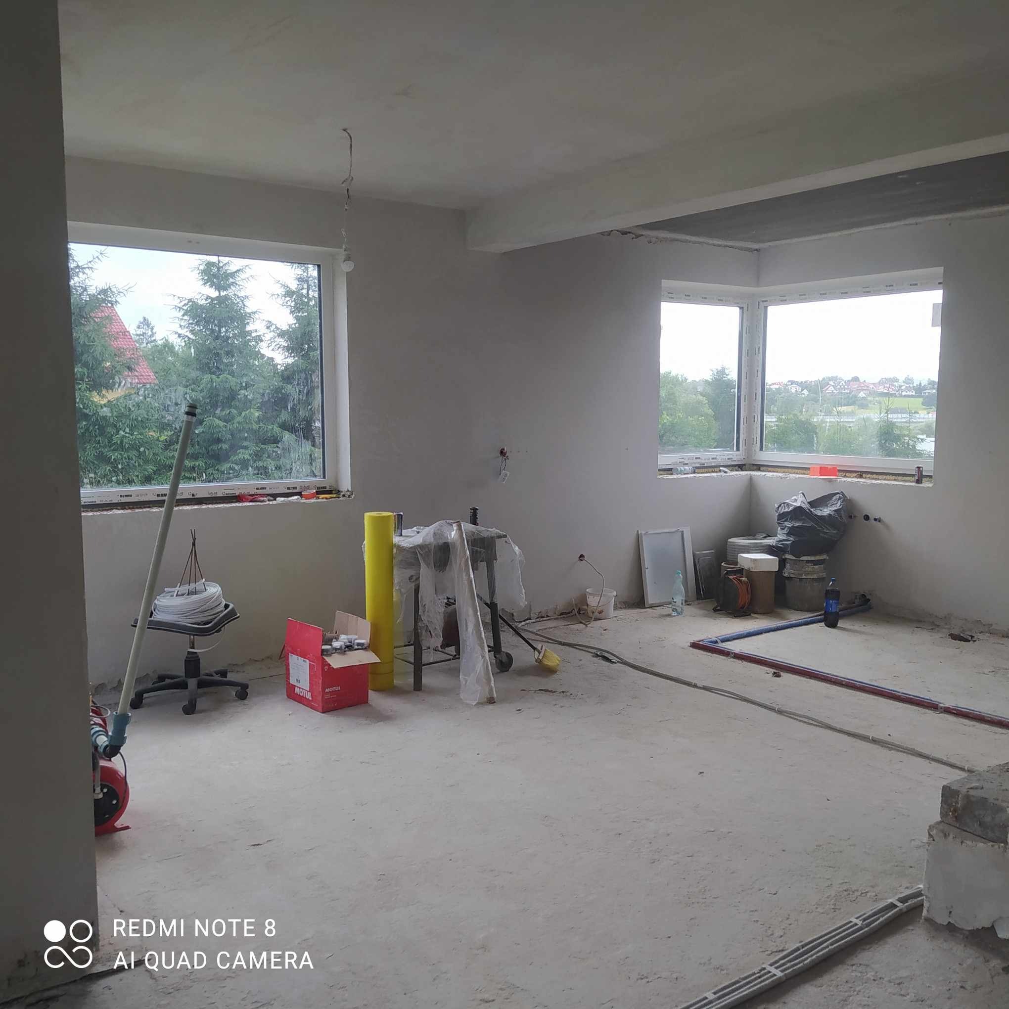 Surowe wnętrze po nałożeniu gładzi gipsowej, widoczne okna z widokiem na zieleń, narzędzia i materiały budowlane rozstawione na podłodze.