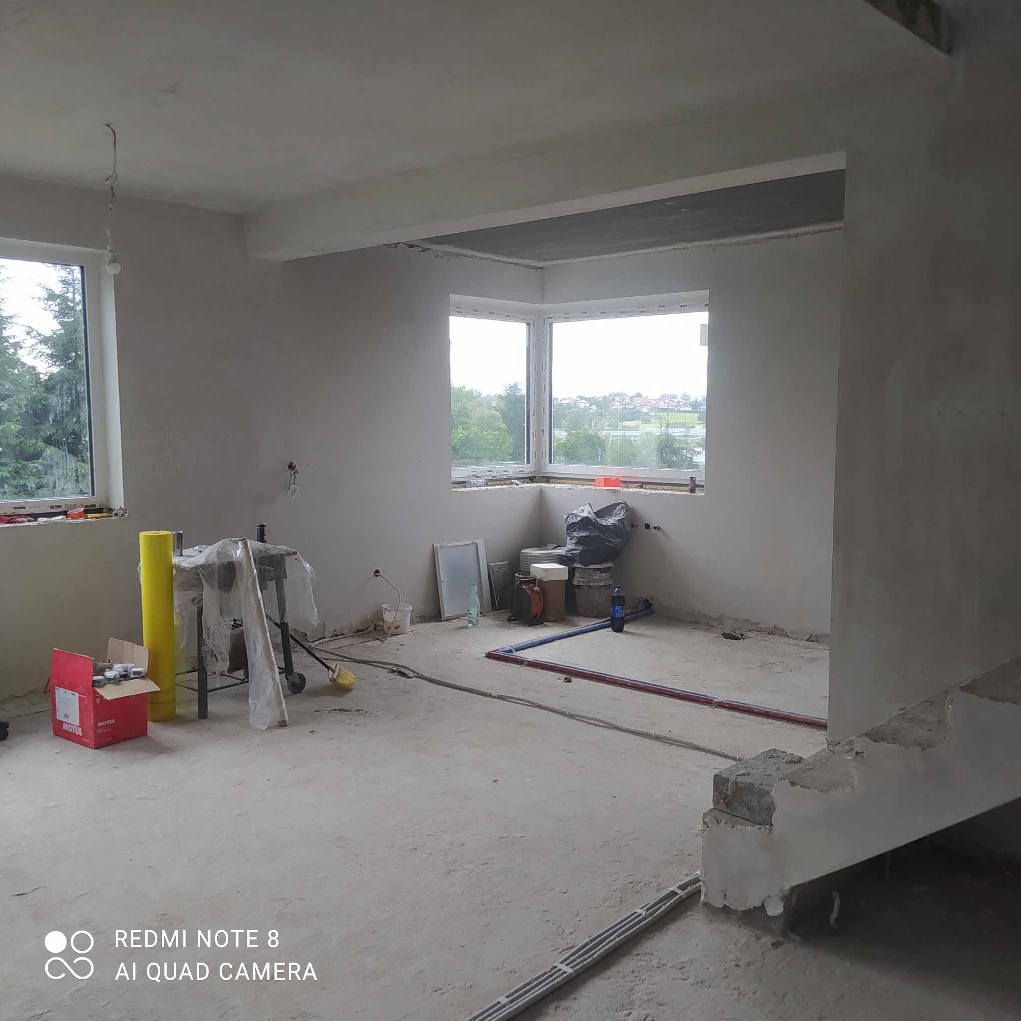 Surowe wnętrze po nałożeniu tynku maszynowego, widoczne okna, instalacje i narzędzia przygotowane do dalszych prac wykończeniowych.
