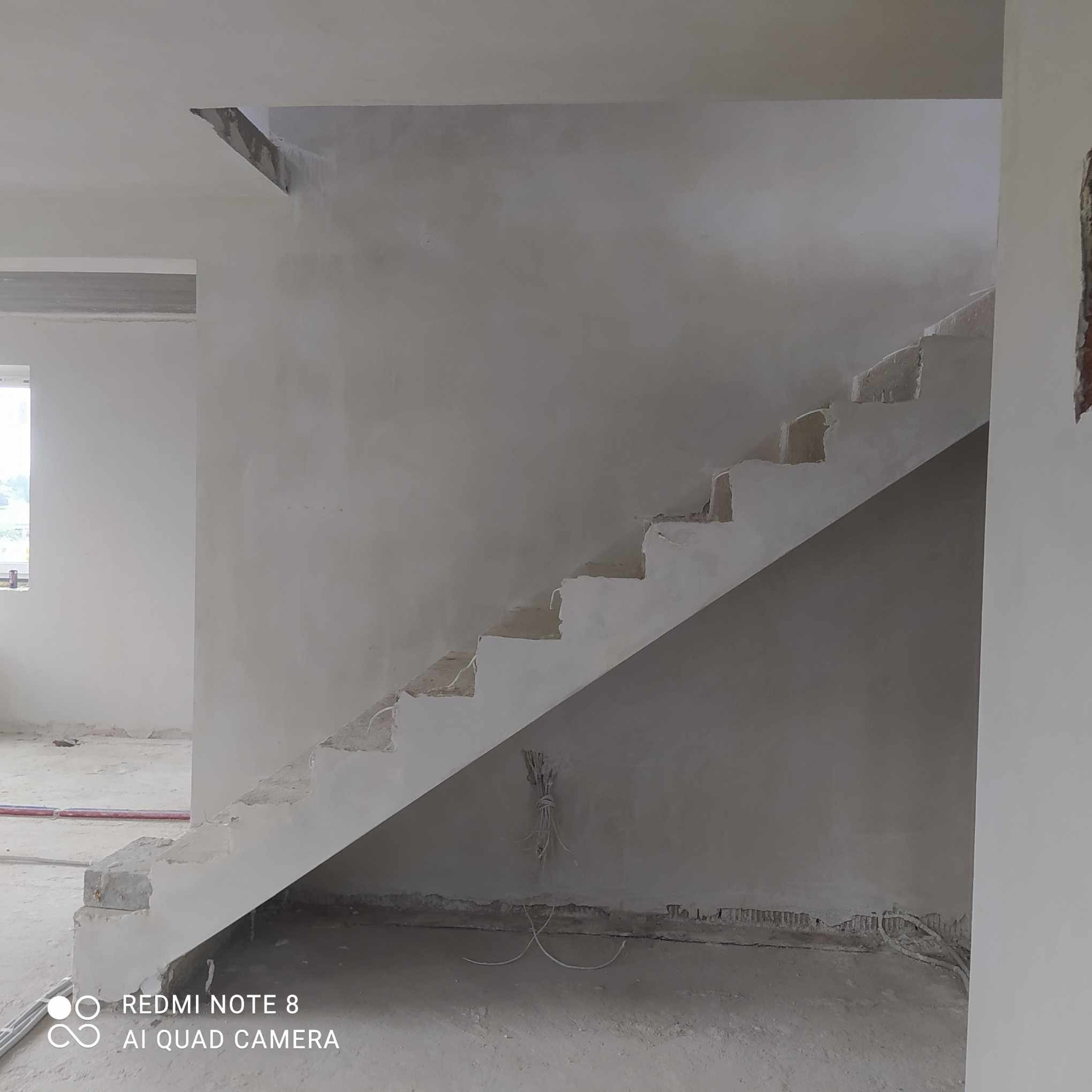 Surowe schody w trakcie prac tynkarskich, widoczne niedoskonałości i wystające przewody elektryczne, szare ściany w stanie deweloperskim.