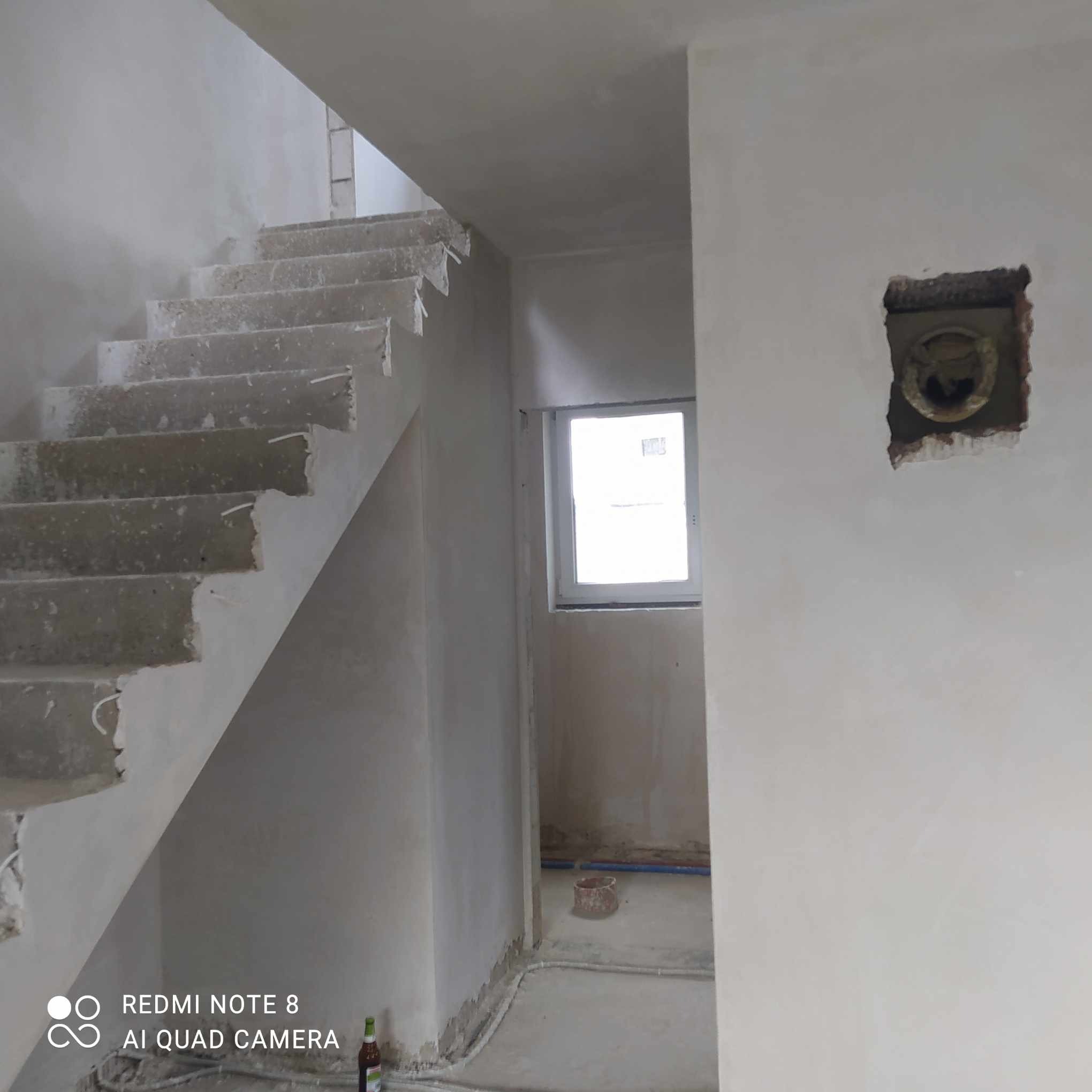 Wnętrze budynku w trakcie prac wykończeniowych: surowe schody betonowe, świeżo otynkowane ściany, otwór instalacyjny w ścianie, widoczne przewody elektryczne i rury, butelka na podłodze.