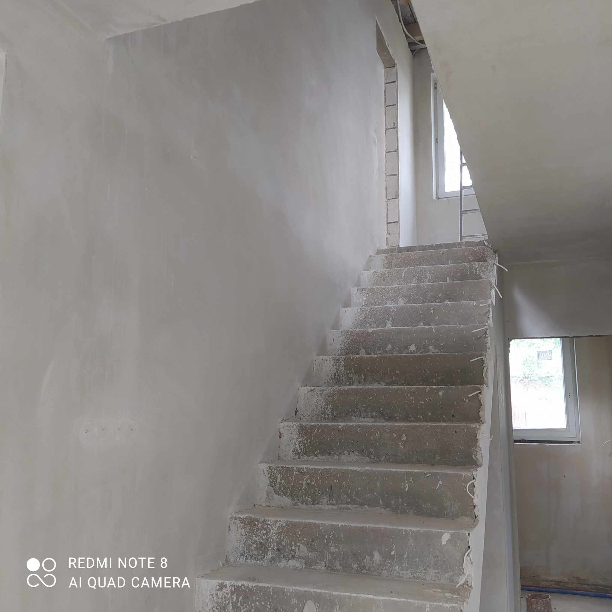 Świeżo otynkowane ściany maszynowo w jasnym odcieniu szarości, surowe betonowe schody z widocznymi niedoskonałościami i fragmentami tynku, okno w tle