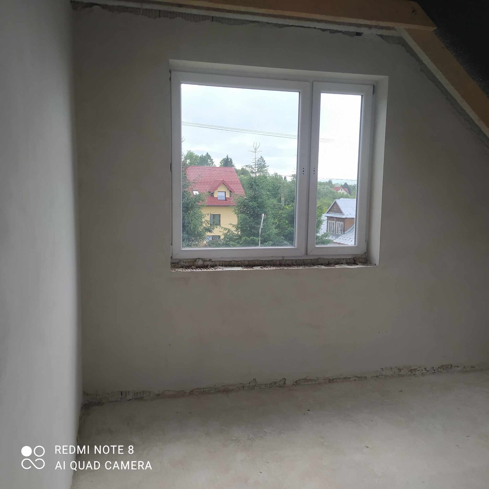 Surowe ściany wewnętrzne po nałożeniu tynku cementowo-wapiennego, widoczne niedoskonałości przy oknie i podłodze, w tle widok na domy przez okno.