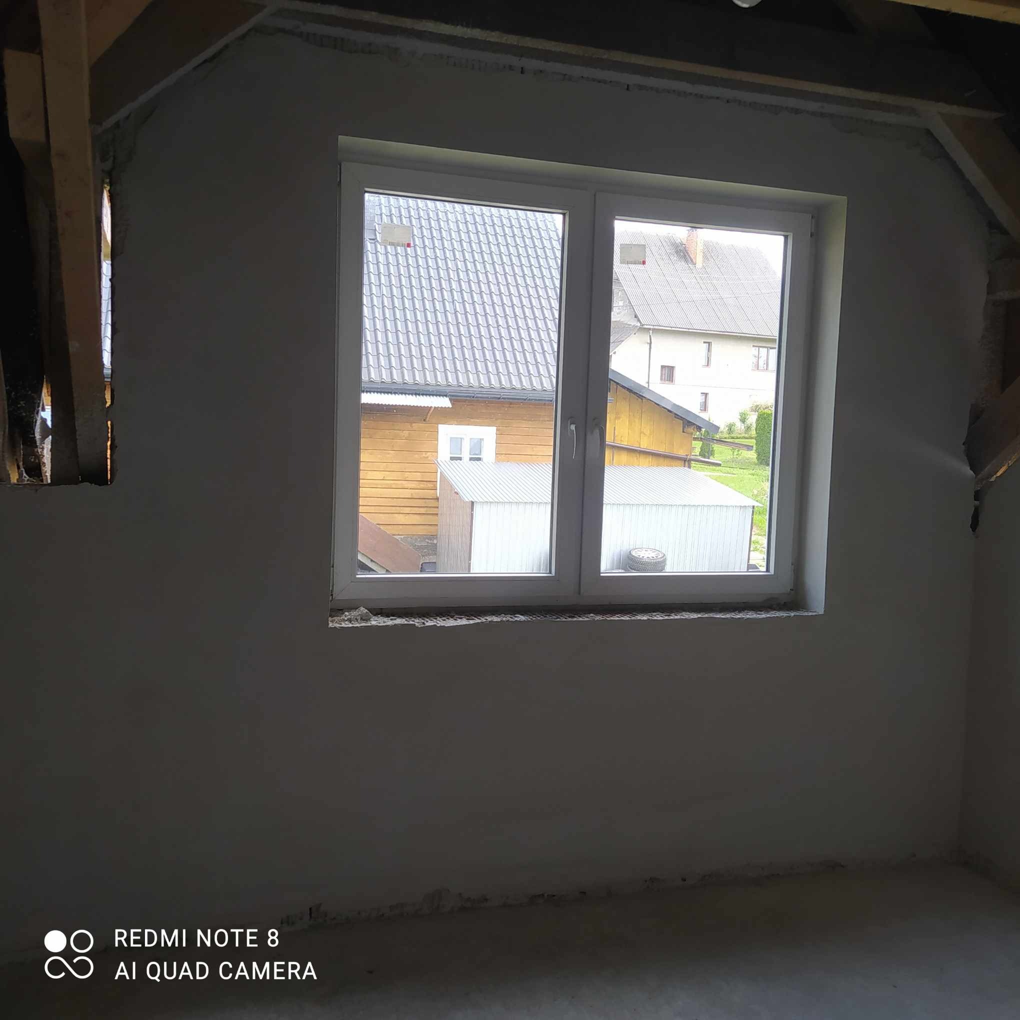 Wnętrze poddasza po nałożeniu tynku, widoczne drewniane elementy konstrukcyjne dachu i okno z widokiem na zabudowania gospodarcze.