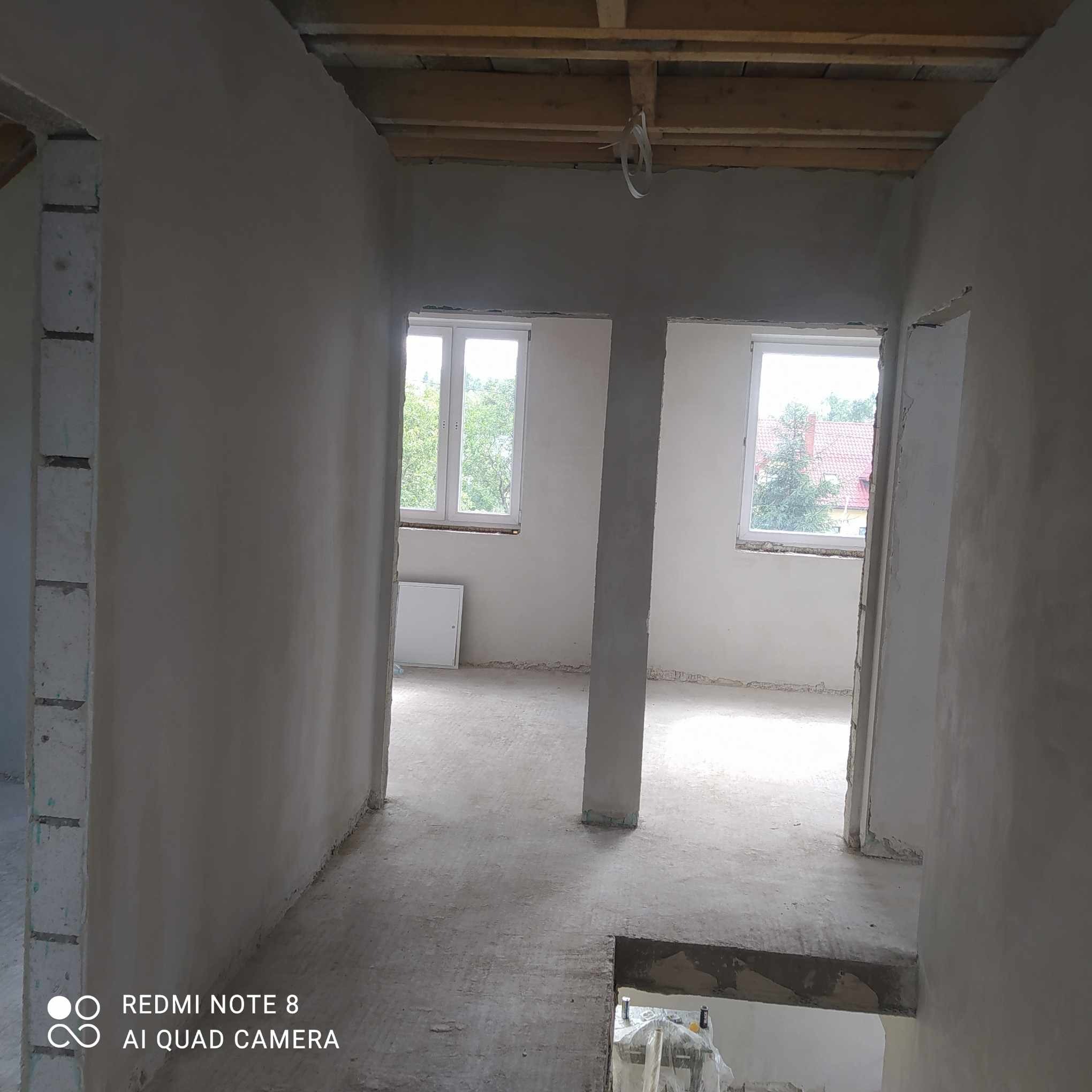 Surowe wnętrze domu po nałożeniu tynków, widoczne ślady prac murarskich, drewniany strop i dwa okna z widokiem na zieleń.