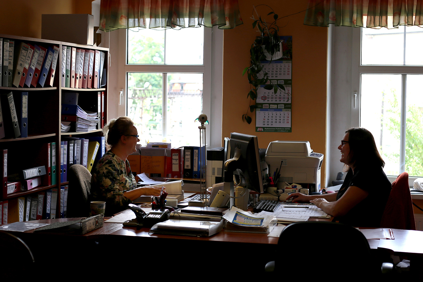 Dwie kobiety pracują w biurze przy biurkach, otoczone segregatorami, komputerami i dokumentami, widoczne kalendarze na ścianie.