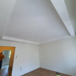 Wnętrze pokoju po remoncie, świeżo pomalowane ściany i sufit z dekoracyjną listwą, widoczny fragment okna i podłogi z paneli, w tle osoba w przejściu do innego pomieszczenia.