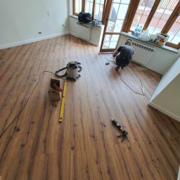 KARO-BUD Sławomir Kadleczek - Montaż paneli podłogowych w pokoju z oknami, widoczny pracownik w trakcie pracy, poziomica, odkurzacz i pistolet do kleju.