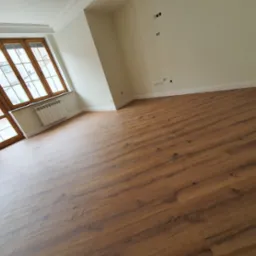 Wykończone wnętrze pokoju z nową podłogą z paneli w odcieniach brązu, jasnymi ścianami z widocznymi gniazdkami i wyłącznikami oraz drewnianą stolarką okienną.