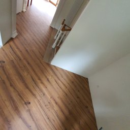 KARO-BUD Sławomir Kadleczek - Nowo ułożone panele podłogowe w korytarzu, widok z góry z perspektywy osoby trzymającej telefon, schody z białymi balustradami w tle.