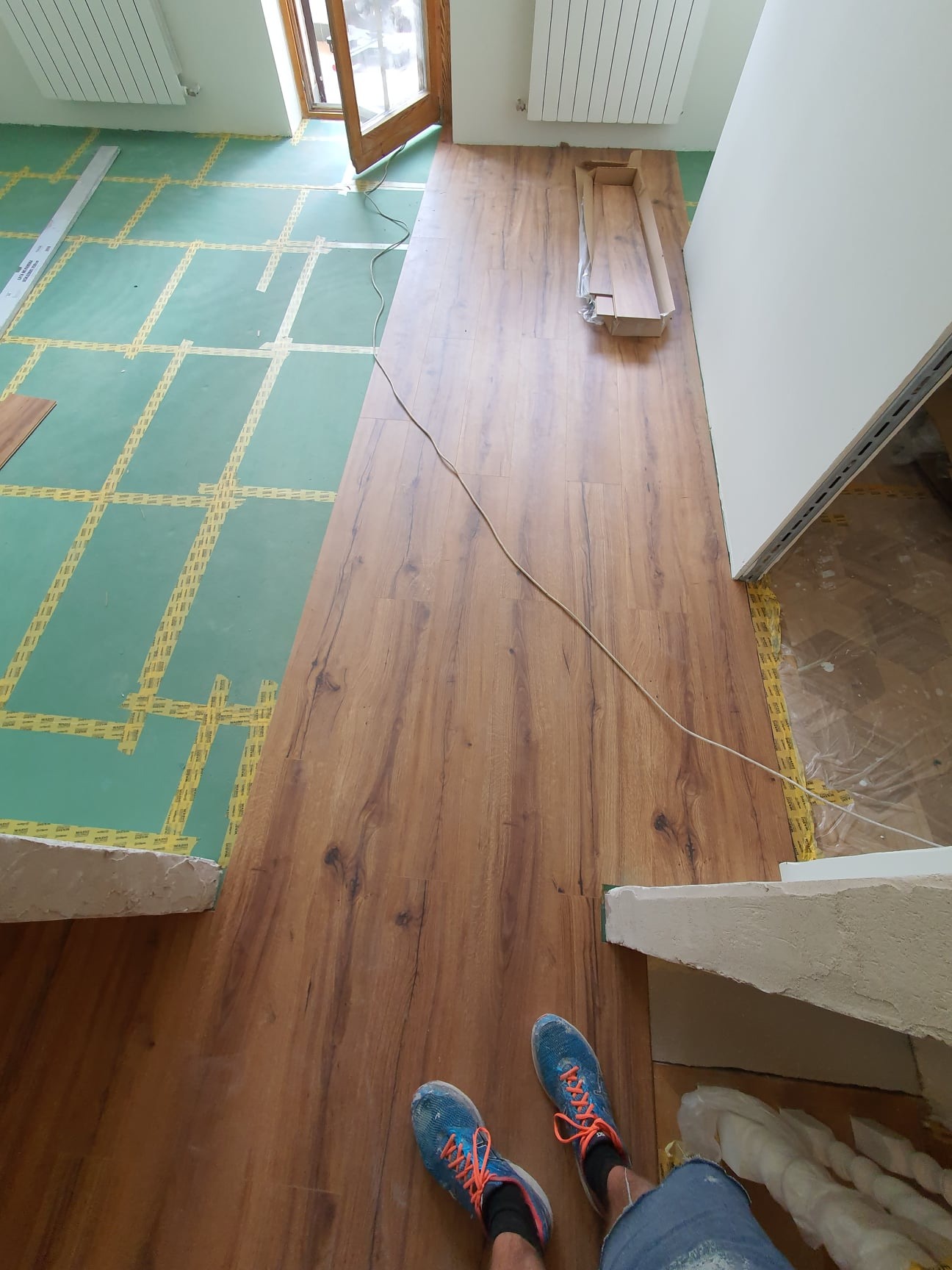 Układanie paneli podłogowych: widok z góry na częściowo ułożoną podłogę z paneli drewnopodobnych, zieloną izolację z żółtą taśmą, oraz niebieskie buty osoby wykonującej pracę.