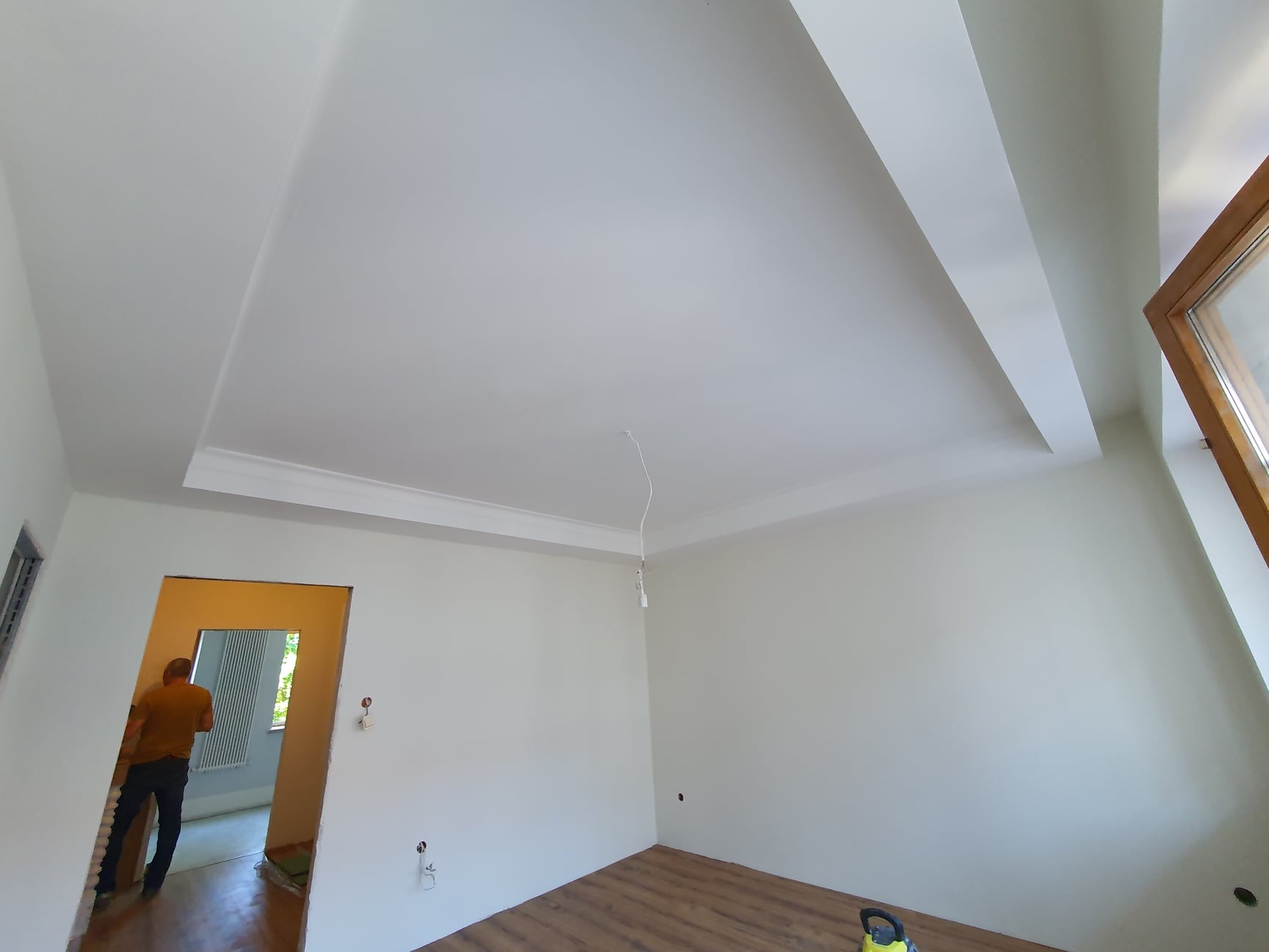 Wnętrze pokoju po remoncie, świeżo pomalowane ściany i sufit z dekoracyjną listwą, widoczny fragment okna i podłogi z paneli, w tle osoba w przejściu do innego pomieszczenia.