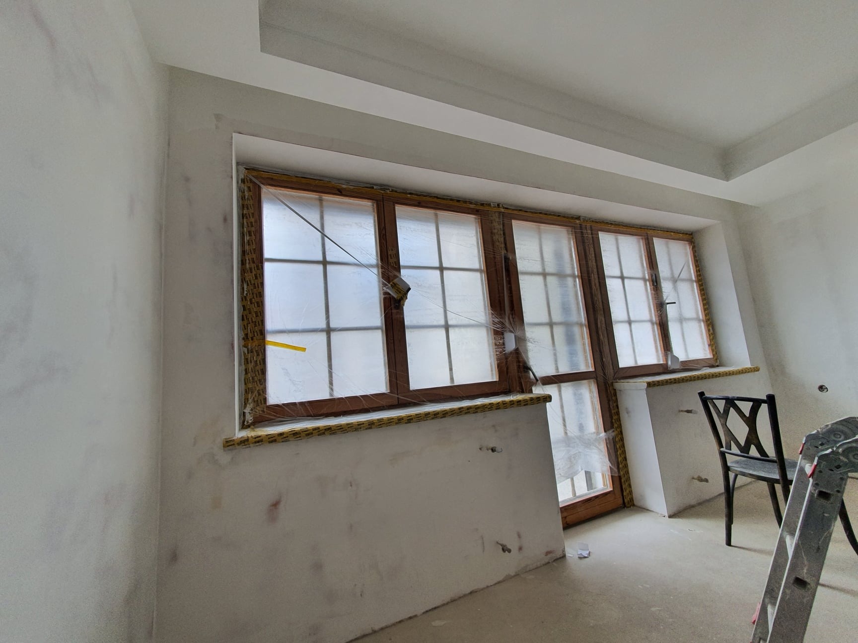 Pokój w trakcie przygotowań do malowania: okna zabezpieczone folią, ściany po szpachlowaniu, widoczne ślady po łatach i nierównościach.