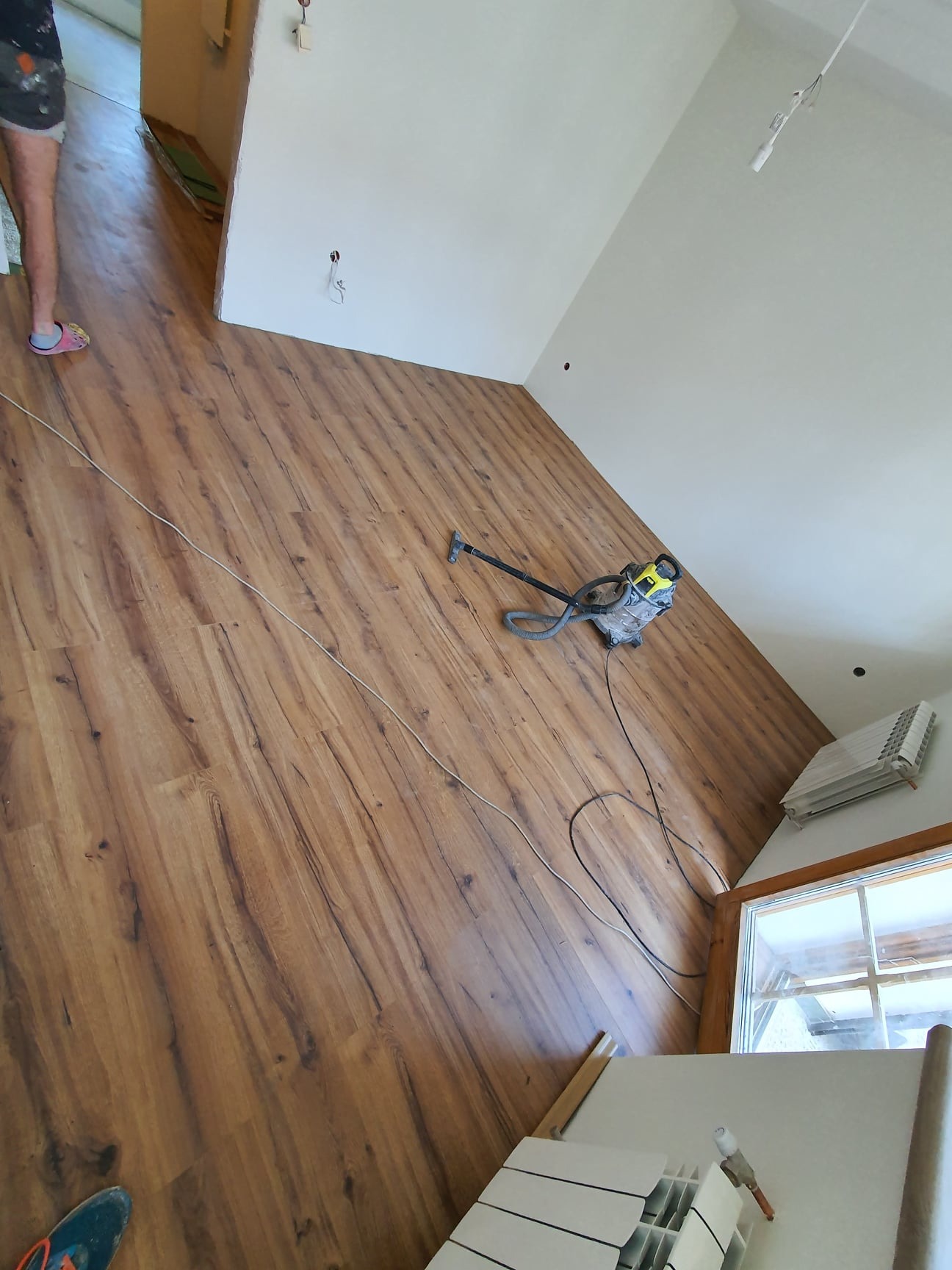 Nowo ułożone panele podłogowe w jasnym pomieszczeniu, z widocznym odkurzaczem przemysłowym do posprzątania po montażu i fragmentem sterty paneli w rogu.