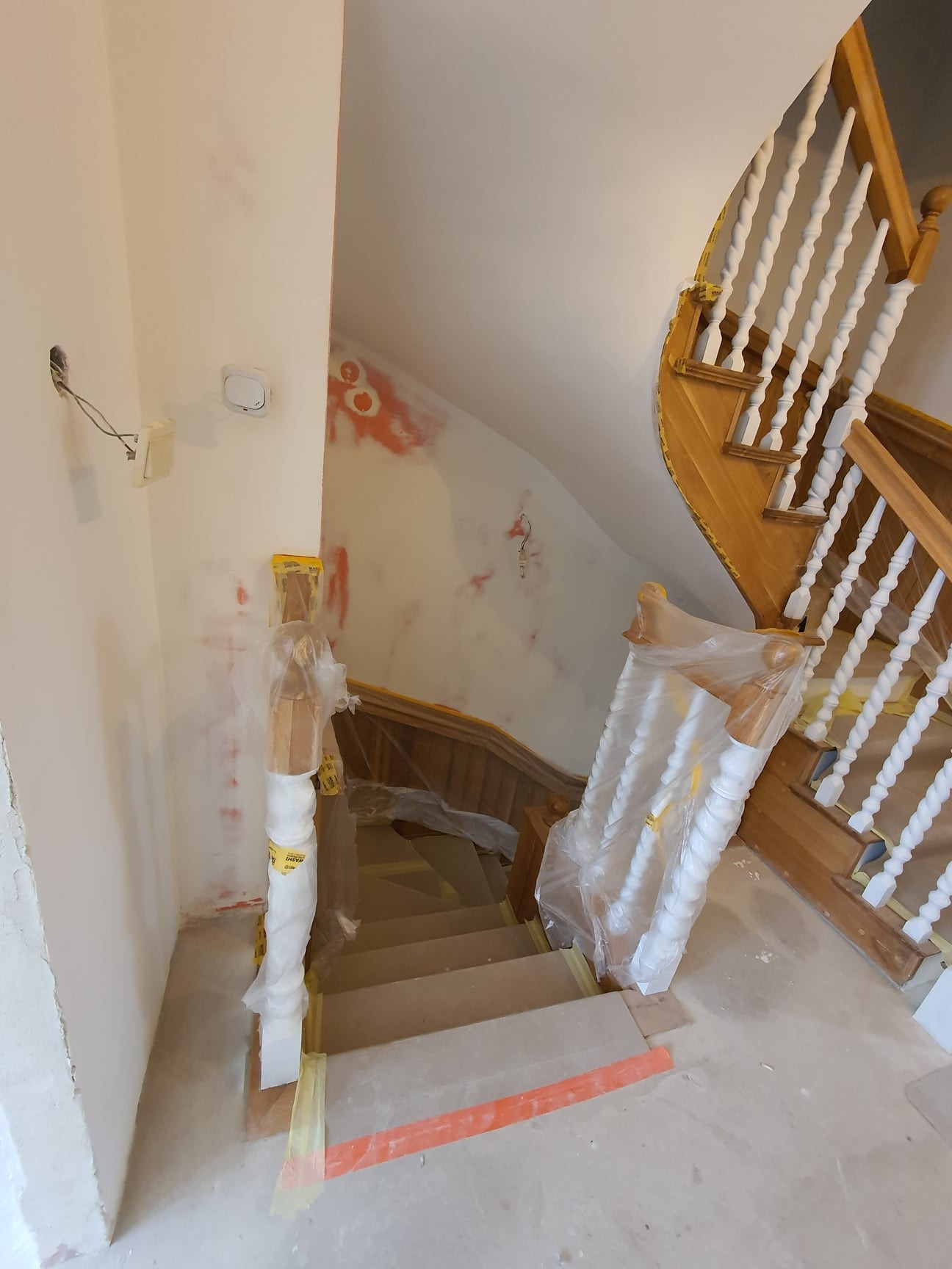 Klatka schodowa w trakcie prac wykończeniowych: zabezpieczone folią balustrady, ściany z widocznymi śladami po szpachlowaniu i wstępnym malowaniu, widoczne instalacje elektryczne.