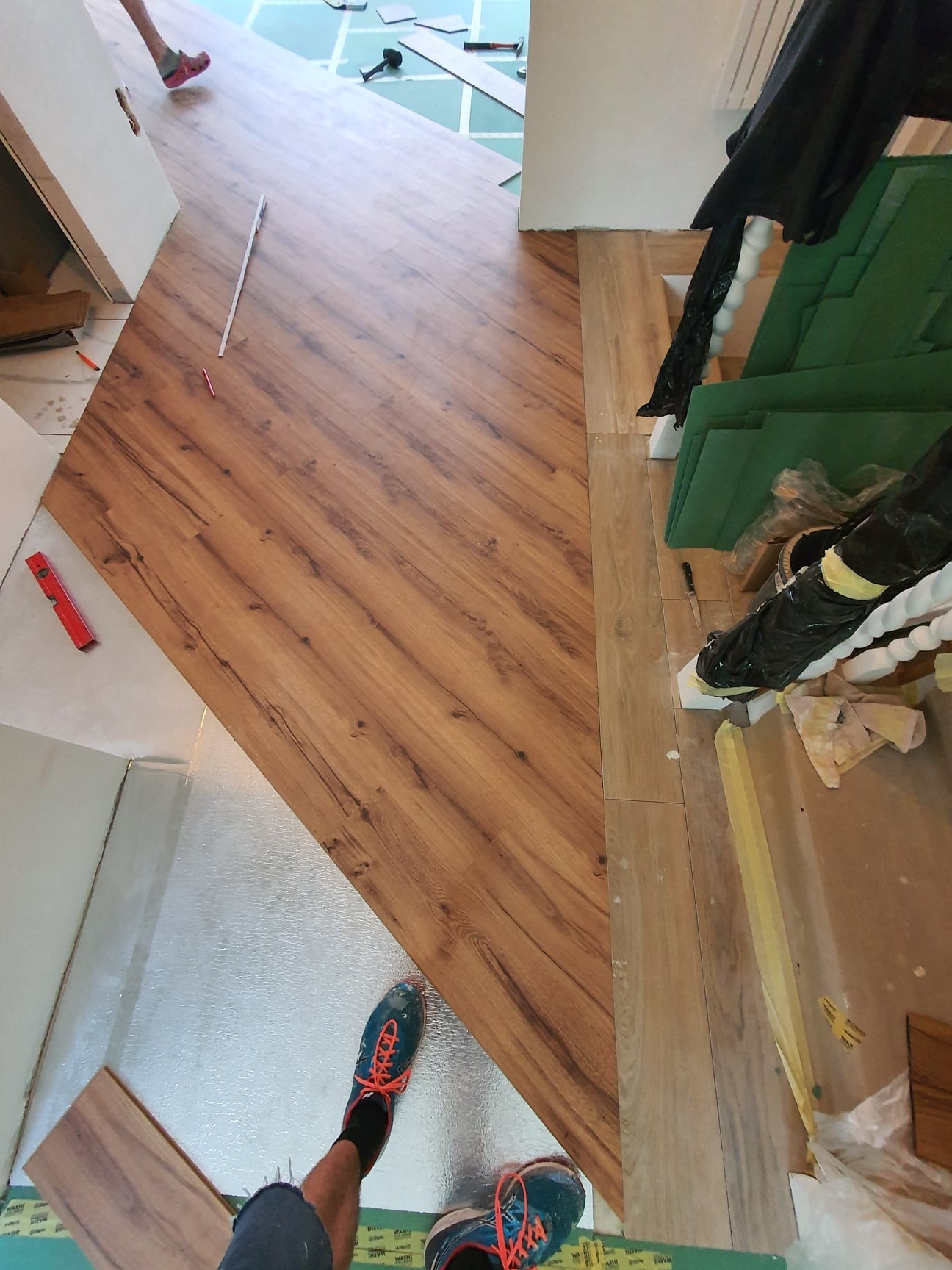 Montaż paneli podłogowych z wyraźnym wzorem drewna, widoczne narzędzia, poziomica, folia izolacyjna i fragmenty butów montera w kadrze.