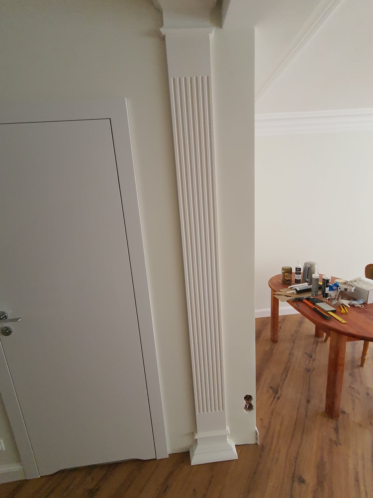 Biała kolumna dekoracyjna z pionowymi żłobieniami, zamontowana przy ścianie obok białych drzwi, z widocznym otworem w ścianie i stołem z narzędziami w tle.