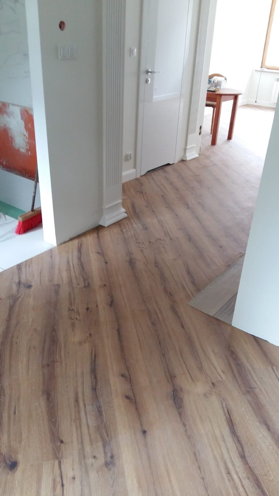 Układanie paneli podłogowych w jasnym wnętrzu, widoczne niedokończone prace przy ścianie i czerwona szczotka.